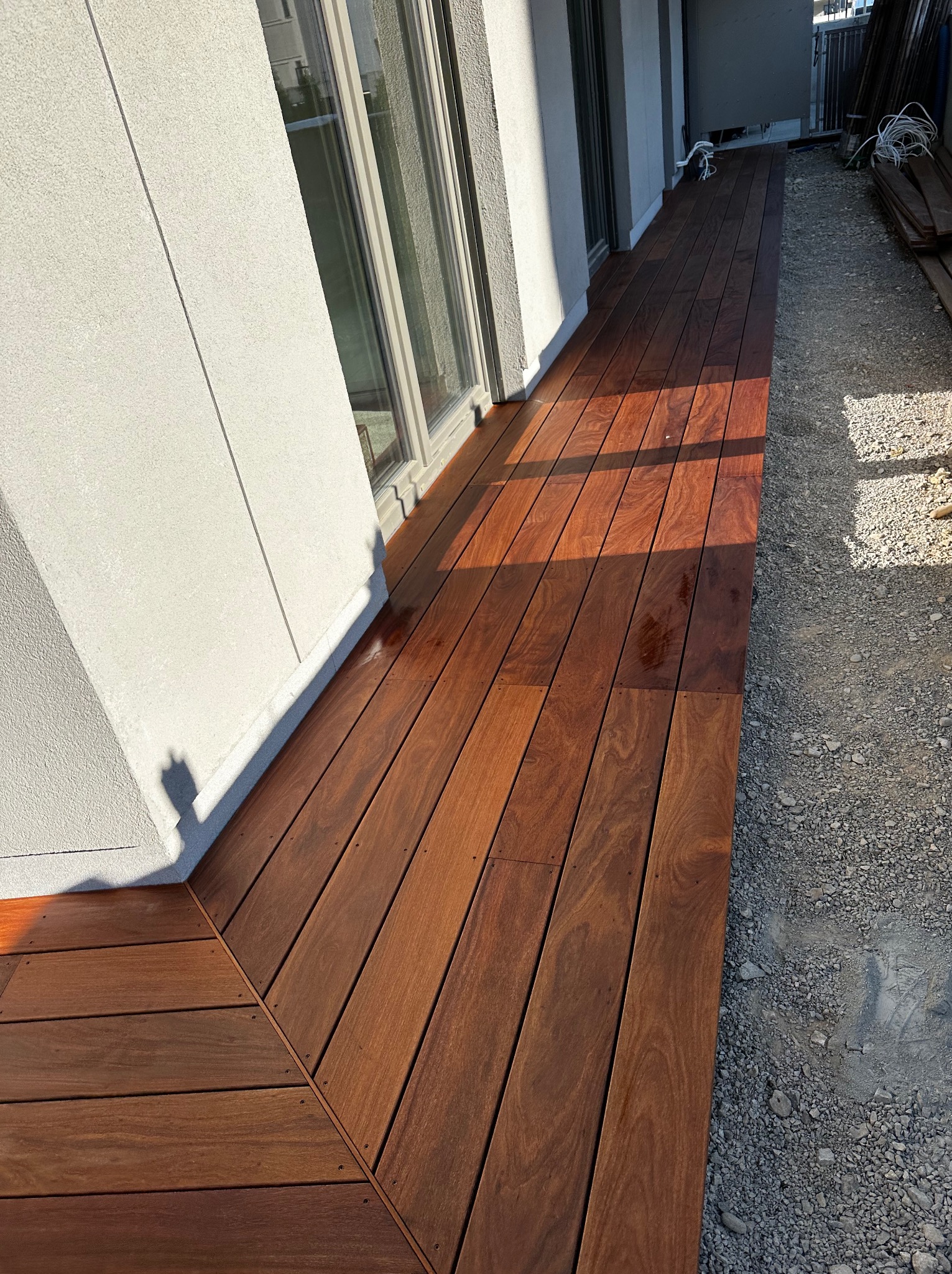 Długi, wąski taras z ciemnego drewna przylegający do budynku z dużymi oknami, wykończony drobnym żwirem. Widoczne ślady po deszczu na desce tarasowej.