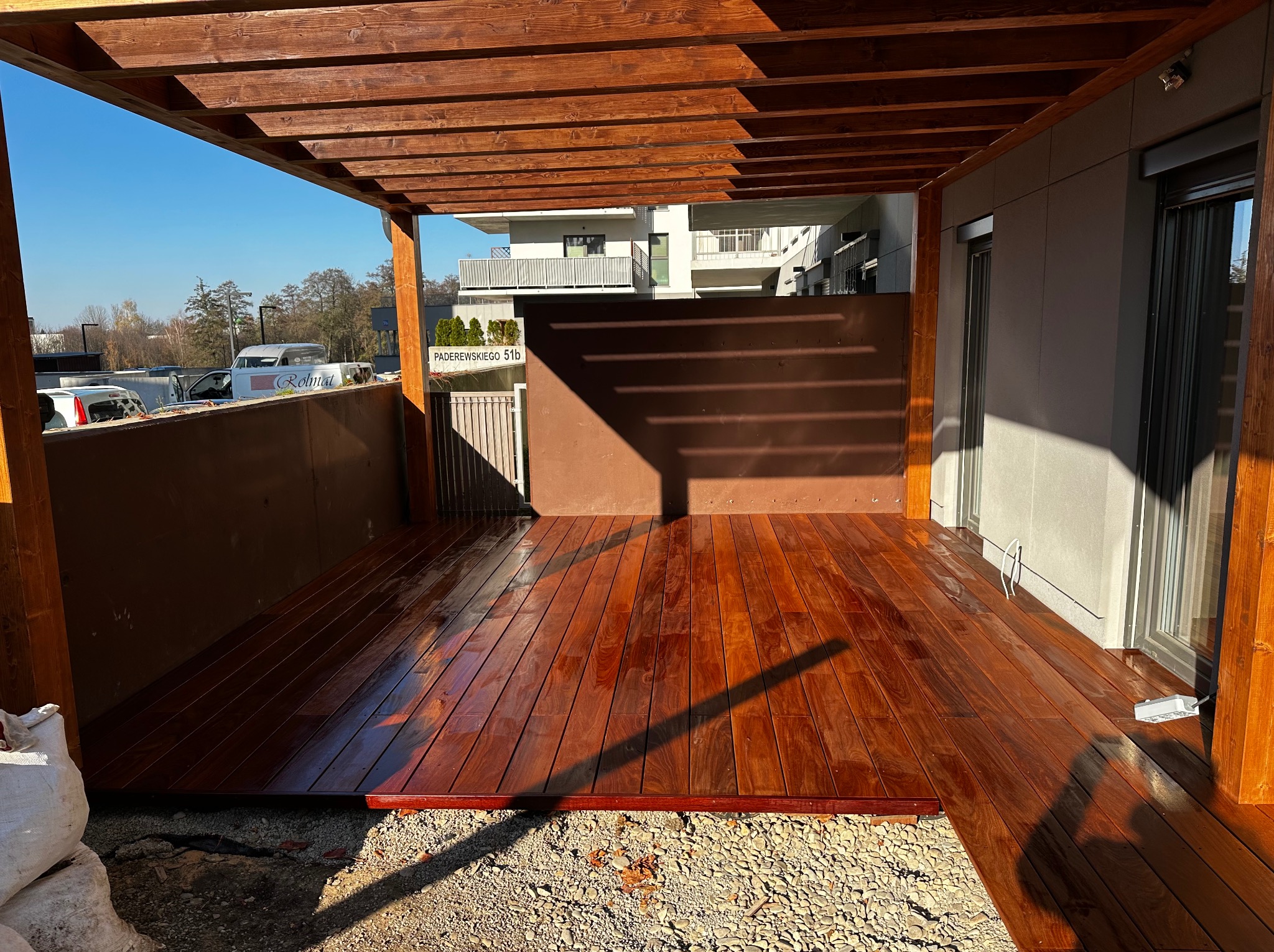 Nowoczesny, drewniany taras z pergolą w ciepłym odcieniu brązu, z widocznym adresem na tabliczce w tle i workiem z materiałem budowlanym w rogu.