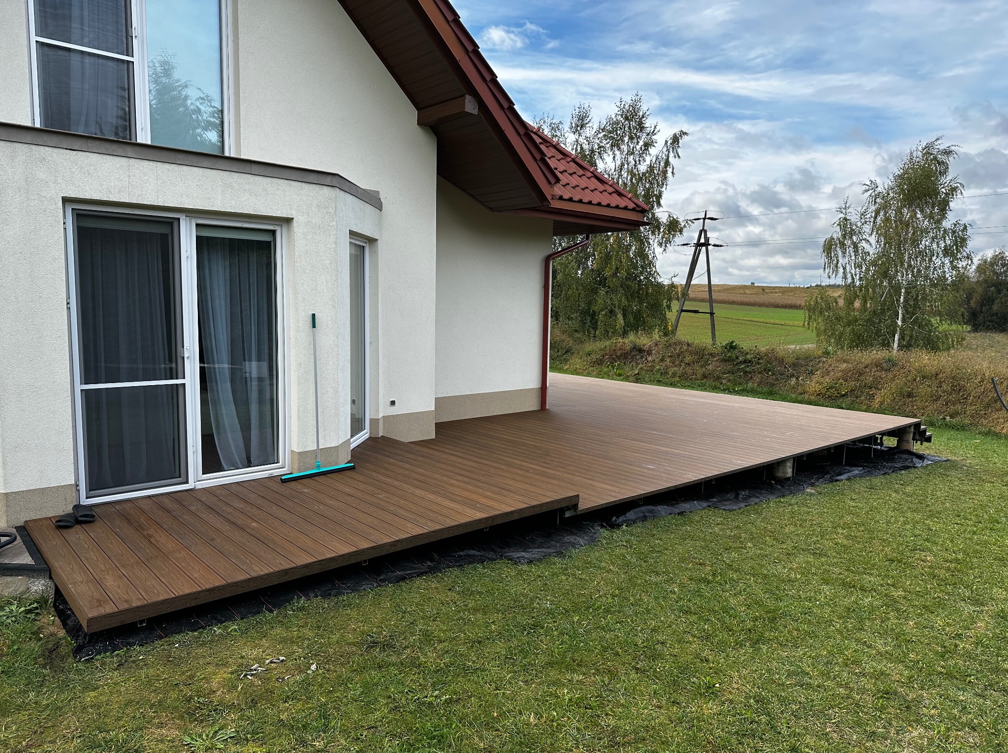 Nowoczesny taras kompozytowy przy domu jednorodzinnym. Brązowe deski tarasowe, widoczne elementy konstrukcyjne. Tło: trawnik, drzewa, błękitne niebo.