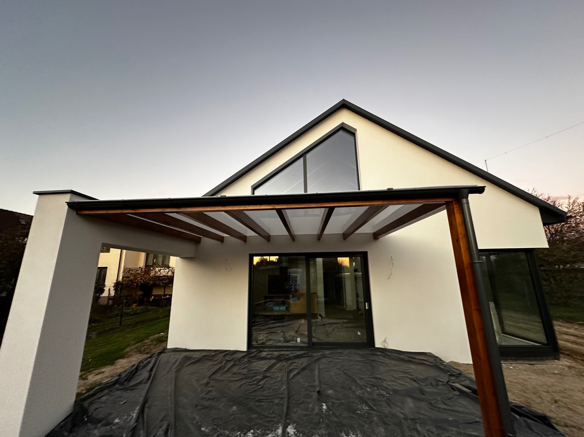 Nowoczesny dom z pergolą o drewnianej konstrukcji, widok z zewnątrz. Dominuje biel ścian i ciemne wykończenia okien. Na ziemi folia ochronna, sugerująca prace wykończeniowe.