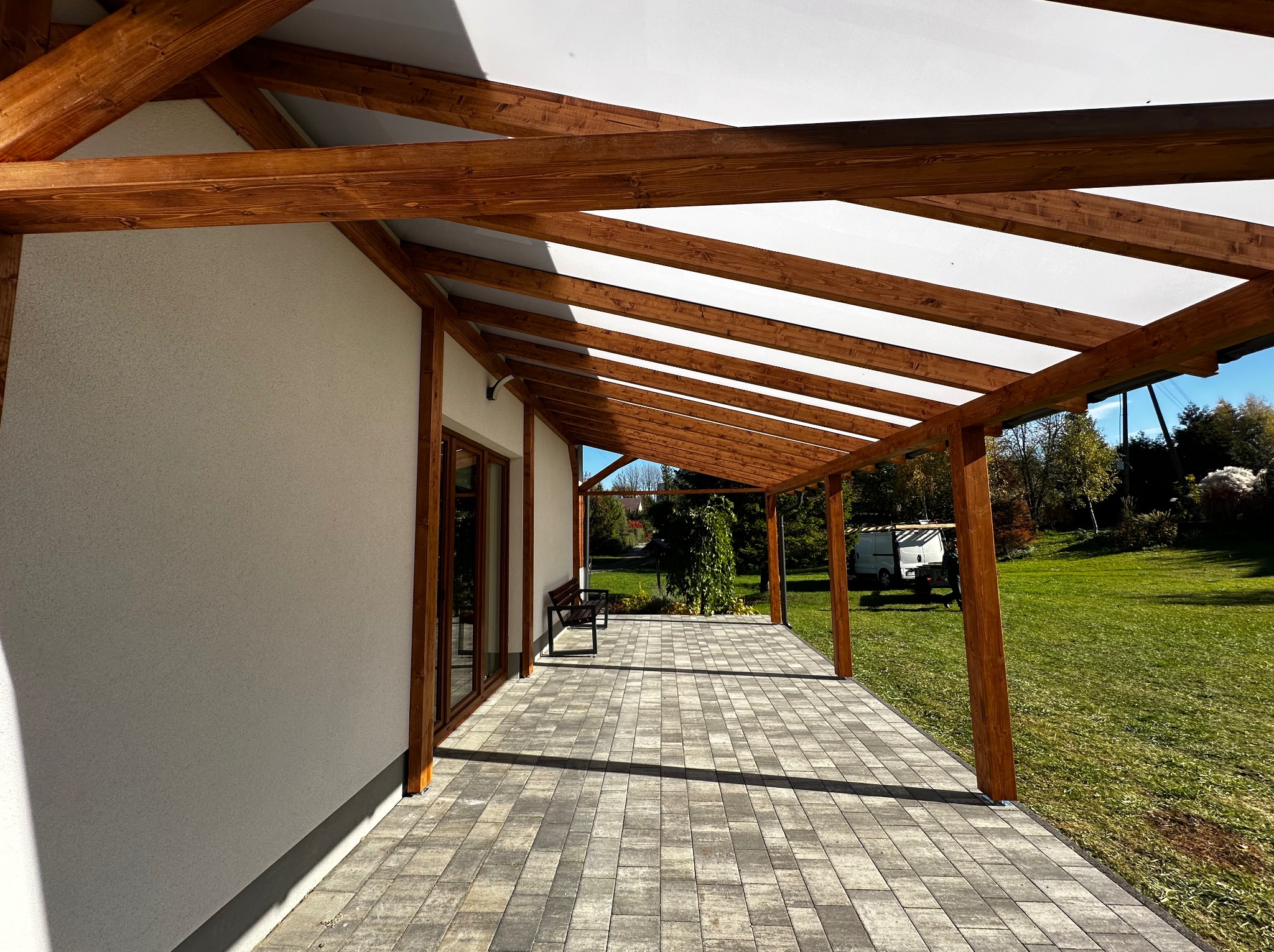 Zadaszony taras z drewnianą konstrukcją i jasną markizą, wyłożony kostką brukową. Widok na ogród i błękitne niebo w tle. Eleganckie połączenie drewna i nowoczesnego designu.