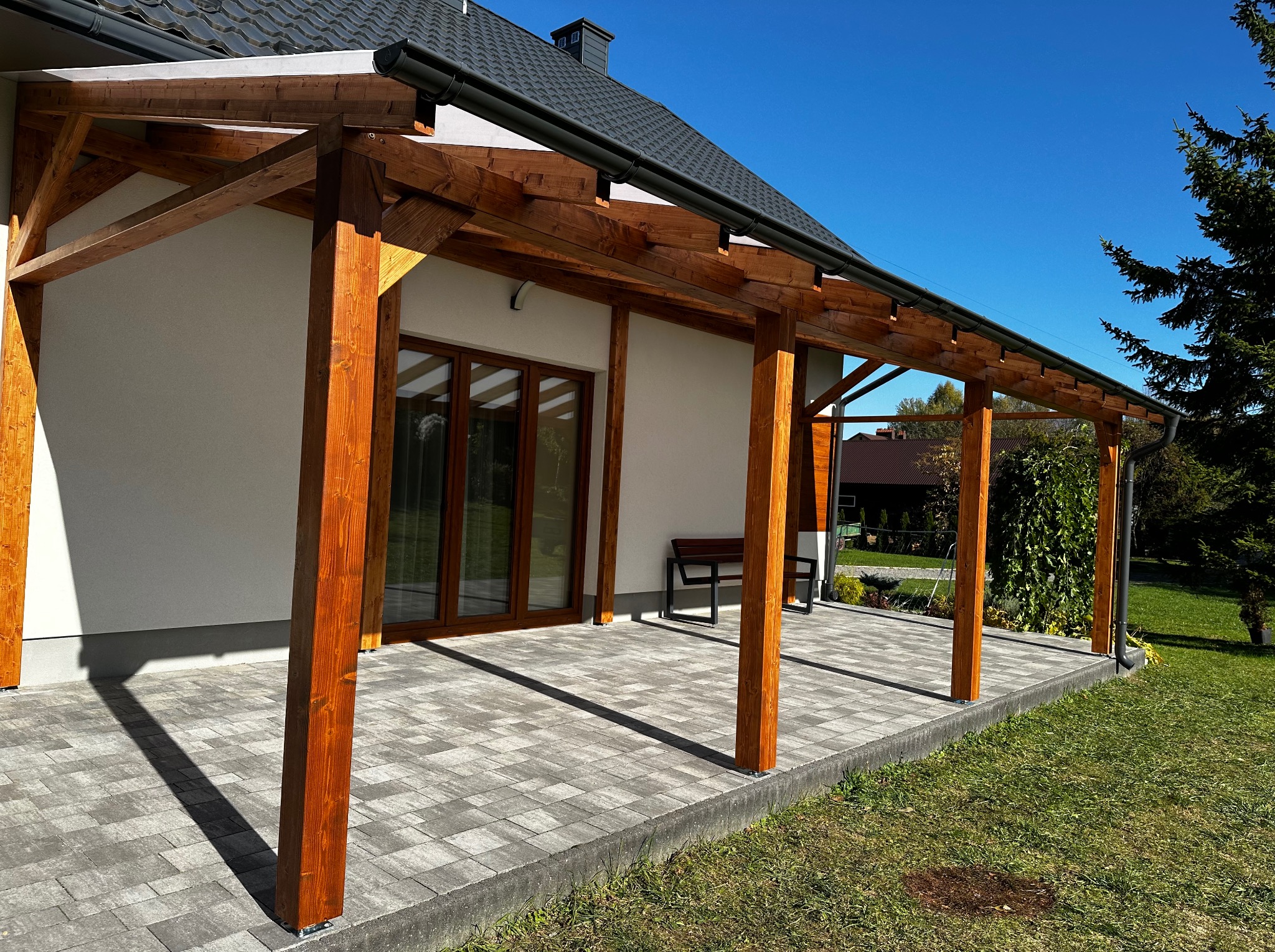 Drewniana pergola zadaszona, z widocznymi elementami konstrukcyjnymi i fragmentem dachu budynku w Terliczce. Na pierwszym planie szara kostka brukowa.