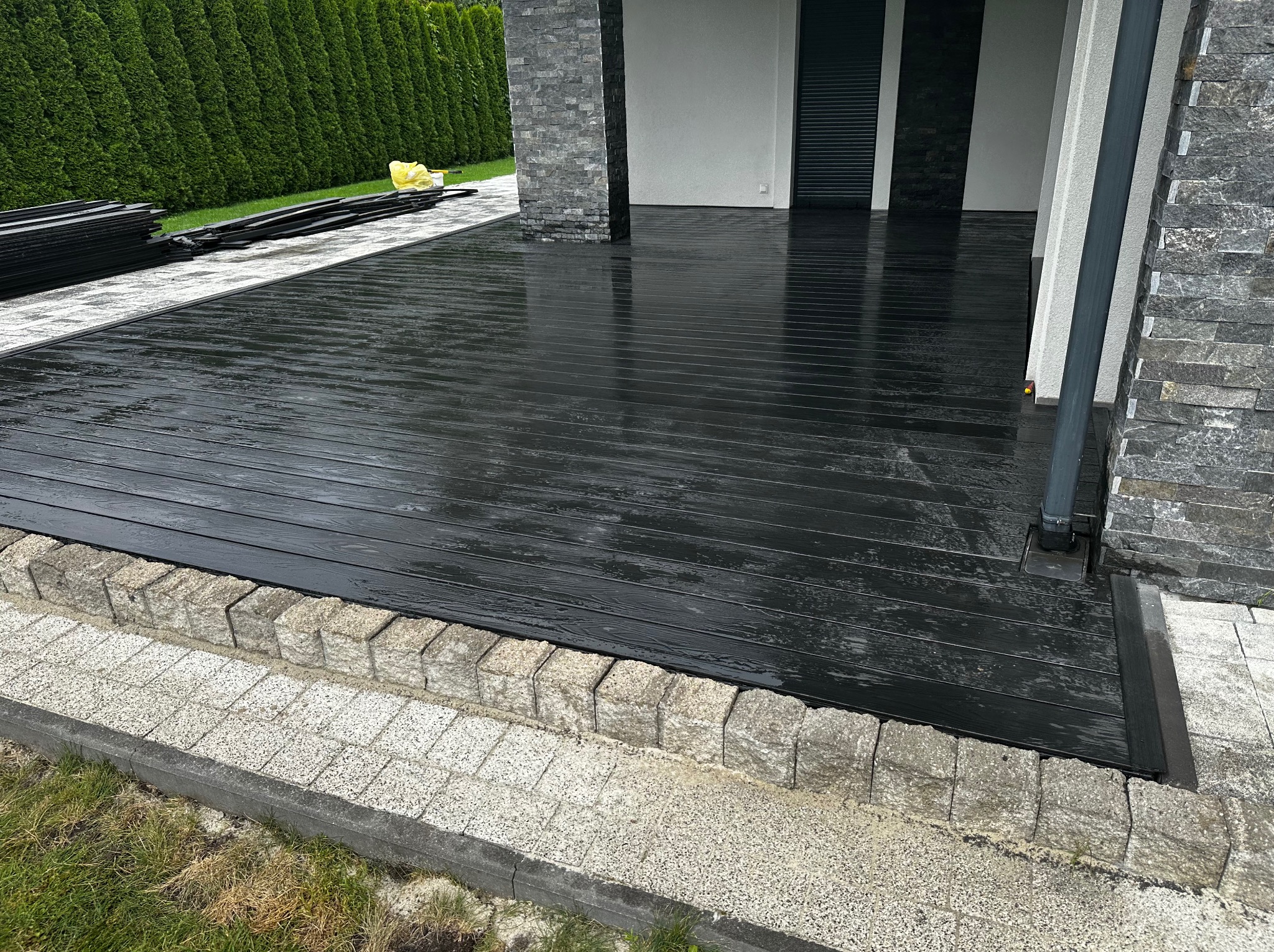 Nowoczesny, czarny taras kompozytowy z desek ułożonych poziomo, obrzeża z kostki granitowej, w tle zielony żywopłot i fragment budynku z kamienną elewacją.