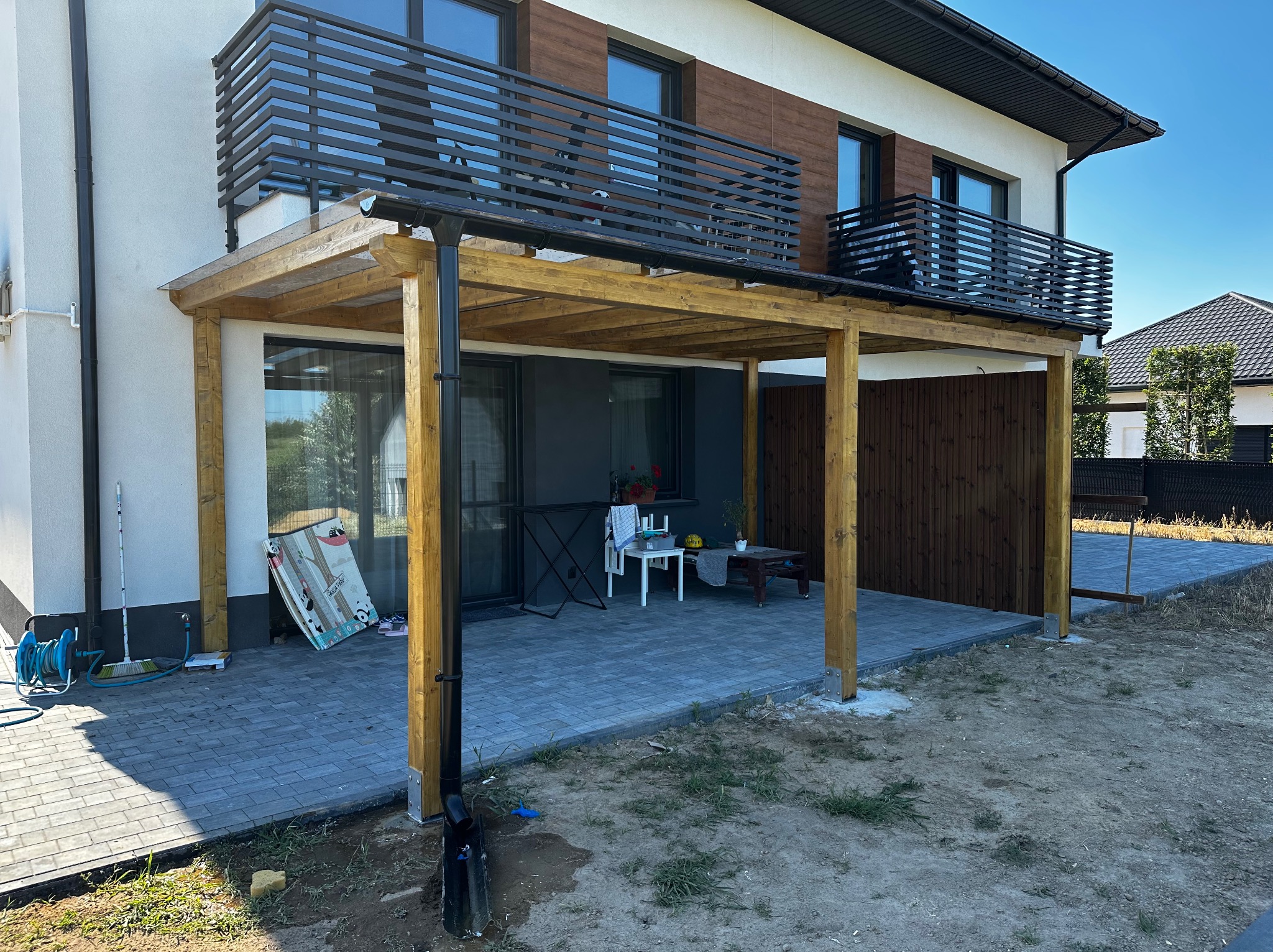 Drewniana pergola zadaszająca taras z szarej kostki brukowej, przylegająca do nowoczesnego domu z balkonem. Widoczny system rynnowy i częściowo niezagospodarowany teren.