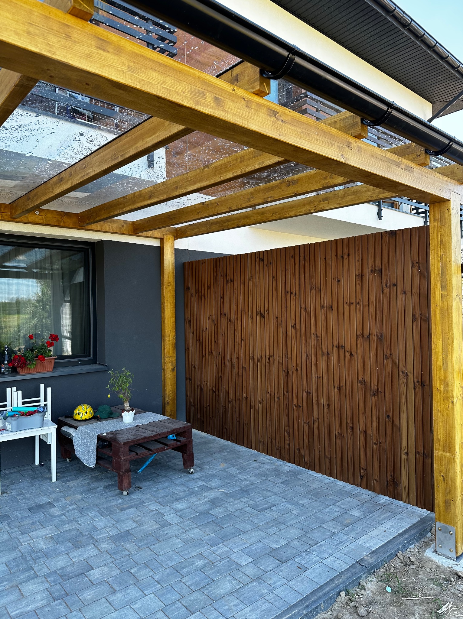 Drewniana pergola z przezroczystym zadaszeniem, przylegająca do domu. Widoczne drewniane panele i meble ogrodowe na wybrukowanym tarasie. Detale: doniczka z kwiatami, żółta czapka.