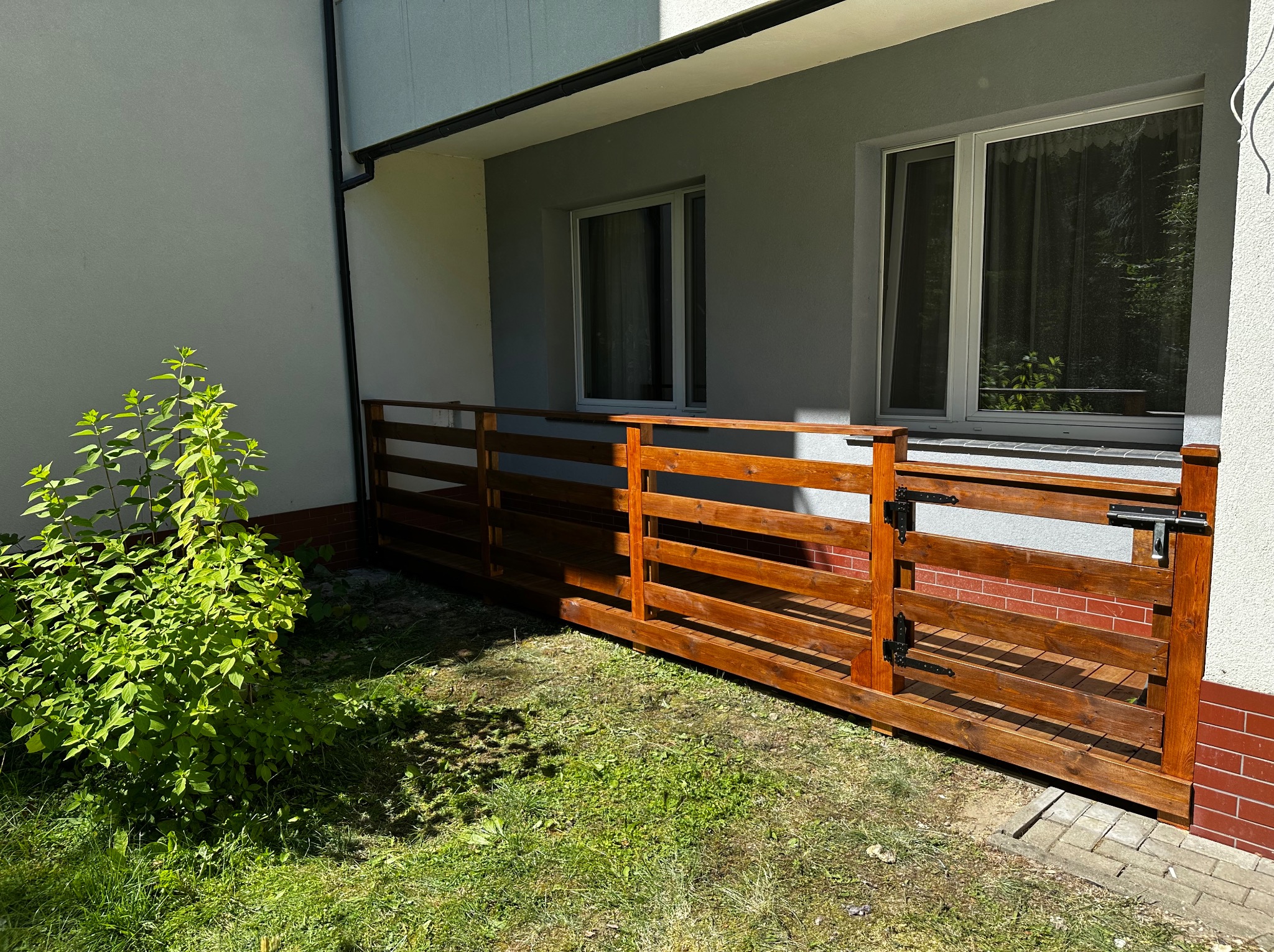 Balkon z drewnianą balustradą i furtką, widok z zewnątrz na trawę i krzew. Balustrada w kolorze ciepłego brązu, z widocznym zamkiem na furtce.