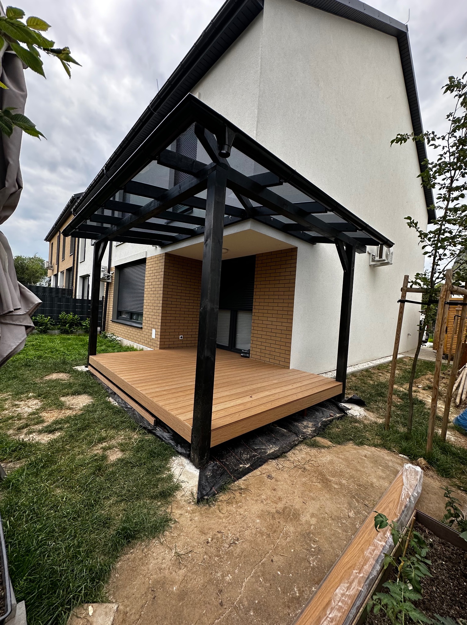 Nowoczesny taras kompozytowy z czarną pergolą przy domu. Jasne deski tarasowe, minimalistyczny design, widok z poziomu gruntu, świeżo posadzona trawa.