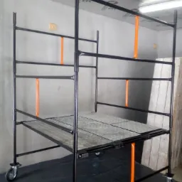 Metalowe rusztowanie budowlane na kółkach z pomarańczowymi elementami mocującymi, ustawione w pomieszczeniu z szarą ścianą i sufitem wykończonym folią paroizolacyjną.