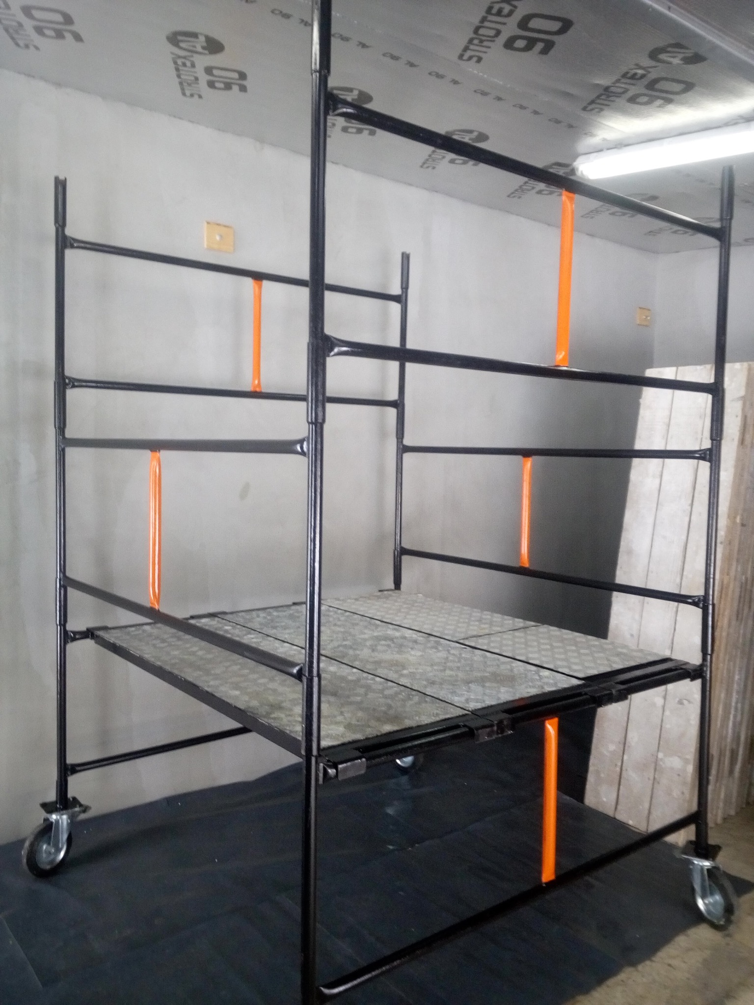 Metalowe rusztowanie budowlane na kółkach z pomarańczowymi elementami mocującymi, ustawione w pomieszczeniu z szarą ścianą i sufitem wykończonym folią paroizolacyjną.