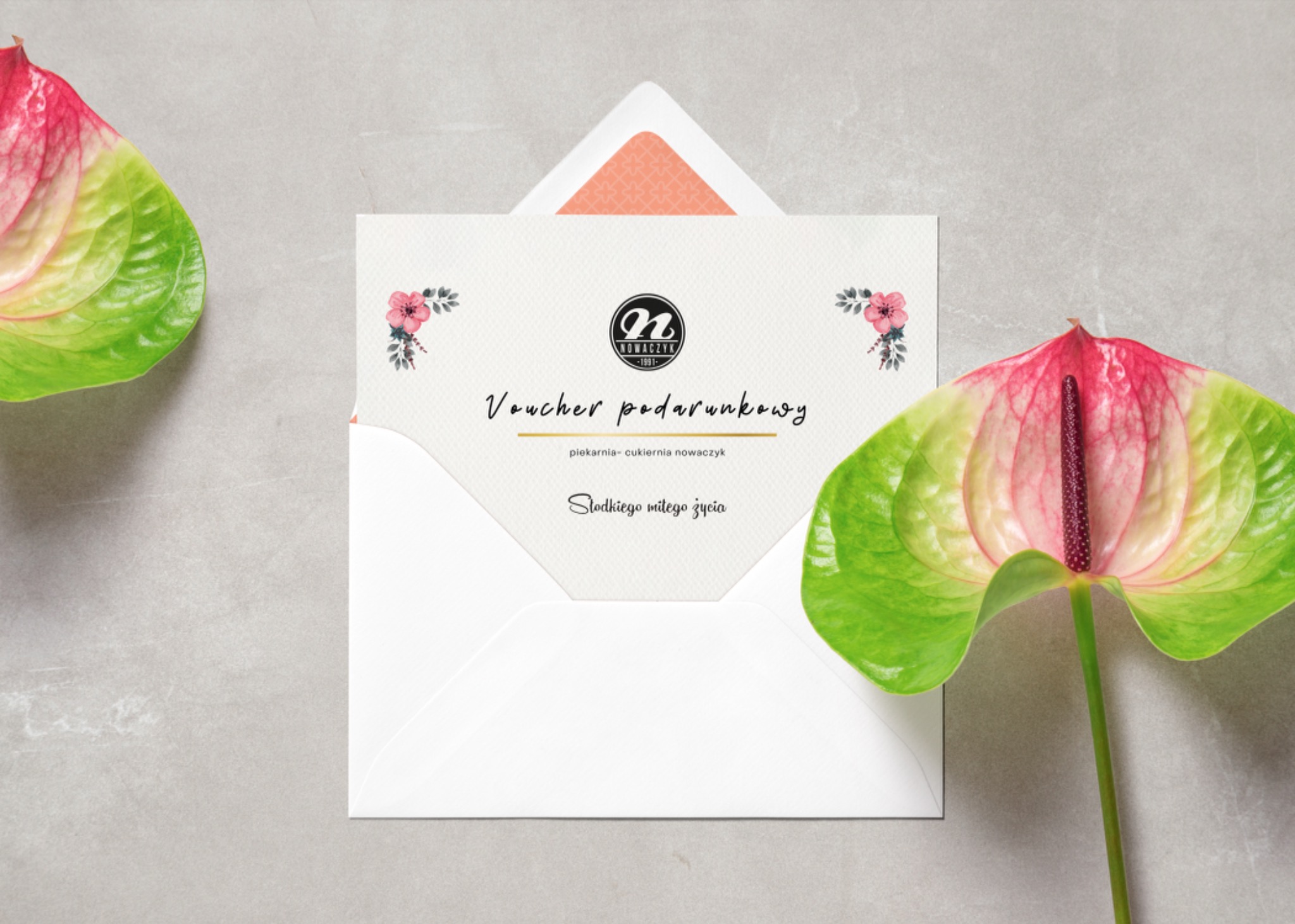 Voucher prezentowy piekarni-cukierni Nowaczyk w białej kopercie, ozdobiony kwiatowymi motywami i anturium w odcieniach zieleni i różu na szarym tle.