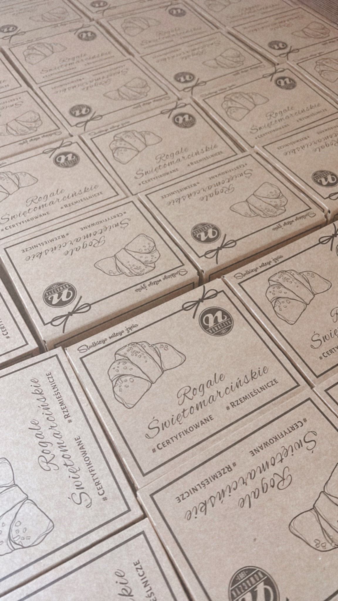 Ułożone rzędy kartonowych pudełek z nadrukowanym rysunkiem rogala, nazwą 'Rogale Świętomarcińskie' i logo firmy piekarniczej.