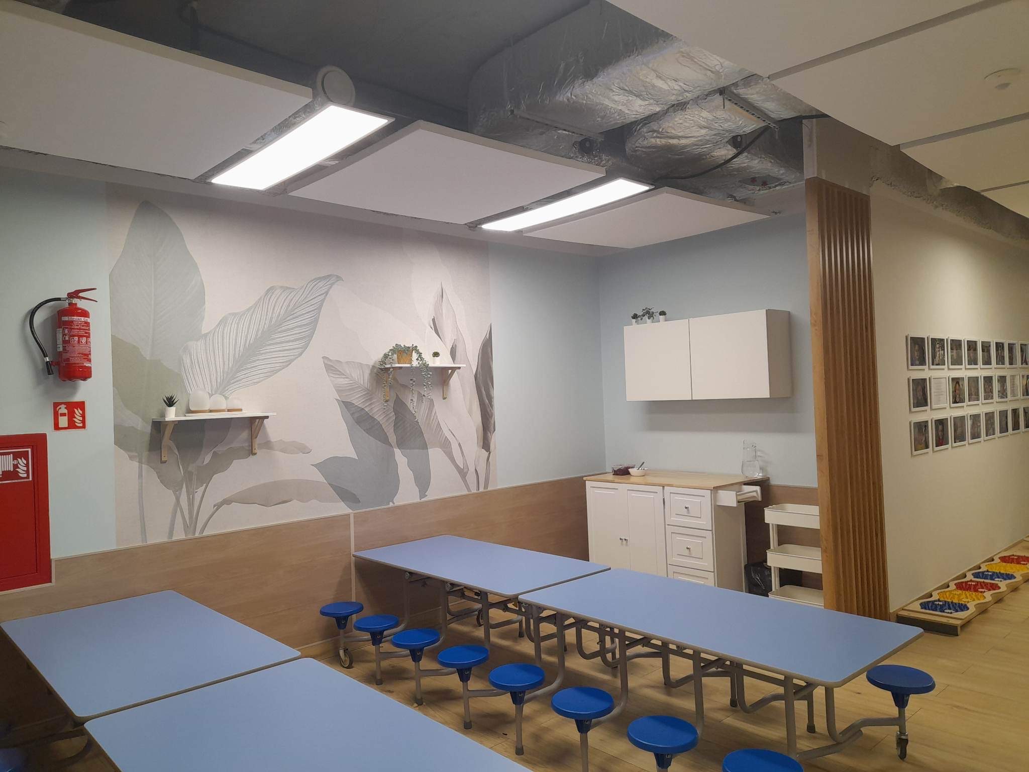 Wnętrze sali zabaw z jasnymi ścianami, wzorem liści na ścianie, niebieskimi stołami i krzesłami, białymi szafkami oraz elementami dekoracyjnymi.