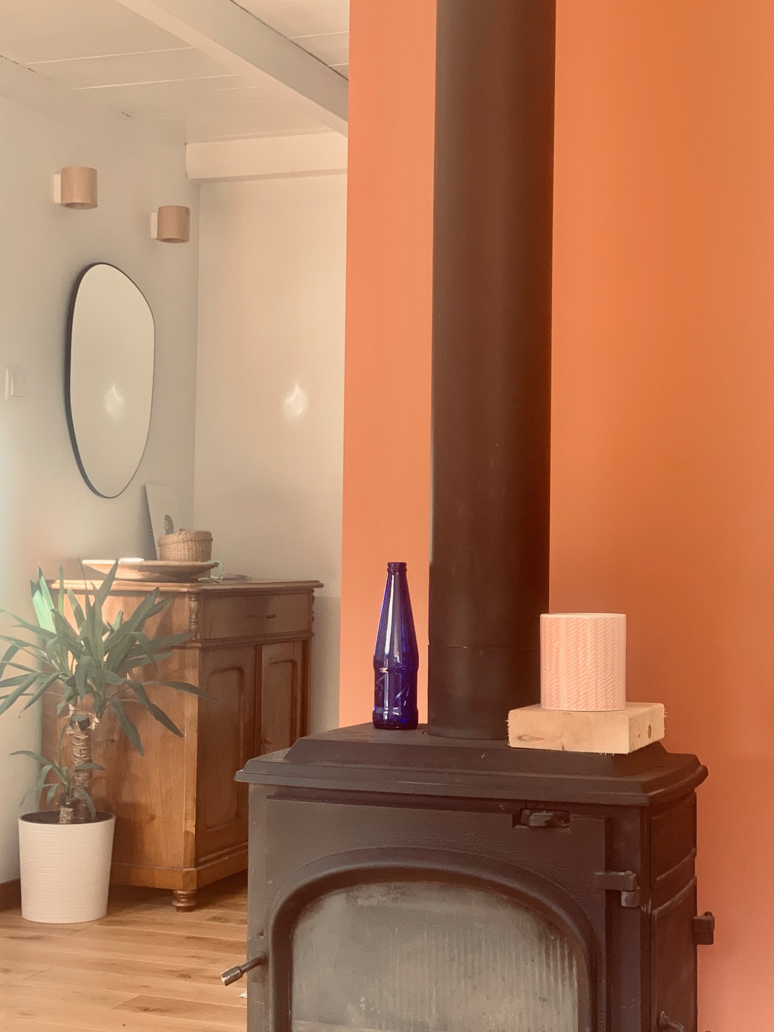 Wnętrze z piecem wolnostojącym, na którym stoi niebieska butelka i pudełko, obok drewniana komoda z lustrem i rośliną w doniczce, pomarańczowa ściana w tle.