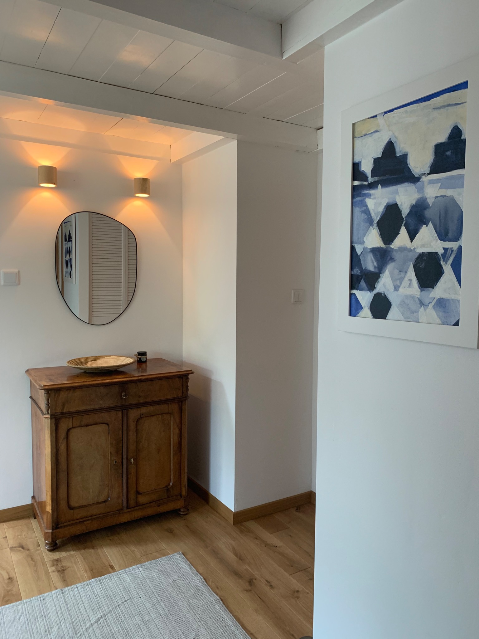 Przedpokój z drewnianą komodą, asymetrycznym lustrem, dwoma kinkietami, obrazem w białej ramie oraz jasnym dywanem na parkiecie.