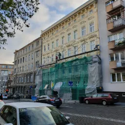 Elewacja kamienicy w trakcie renowacji, widoczne rusztowania i siatka ochronna, pracownicy na rusztowaniu, częściowo odnowiona fasada w jasnym kolorze, ulica z zaparkowanymi samochodami...