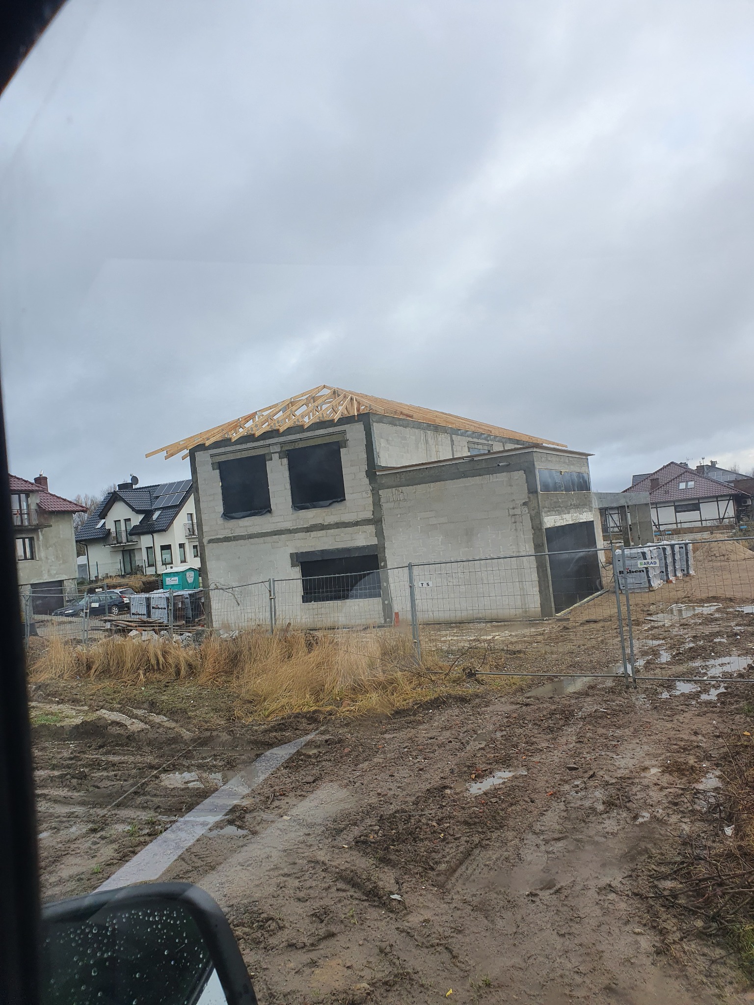 Surowy stan domu jednorodzinnego w Szczecinie z widoczną konstrukcją dachu i niezabezpieczonymi otworami okiennymi, otoczonego błotnistym terenem budowy i tymczasowym ogrodzeniem.