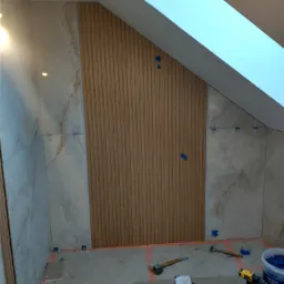 Poddasze w trakcie prac wykończeniowych, ściany częściowo pokryte płytkami imitującymi marmur oraz panelami ściennymi, na podłodze narzędzia glazurnicze i wiadro z klejem, widoczne krzyżyki...