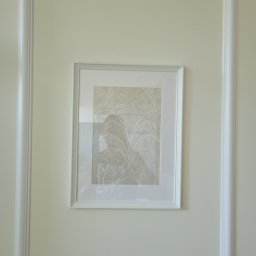 Remodern Filip Biały - Ściana w kolorze złamanej bieli z dekoracyjną ramą i zawieszonym obrazem w białej ramce, przedstawiającym abstrakcyjny wzór w odcieniach beżu i szarości.