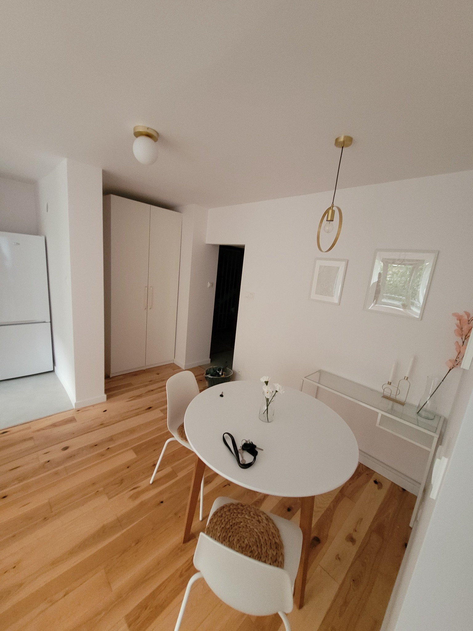 Jasny pokój z drewnianą podłogą, białymi ścianami i okrągłym stołem z bukietem białych kwiatów. Widoczne białe krzesła z plecionymi siedziskami, biała komoda, minimalistyczne obrazy na ścianie...