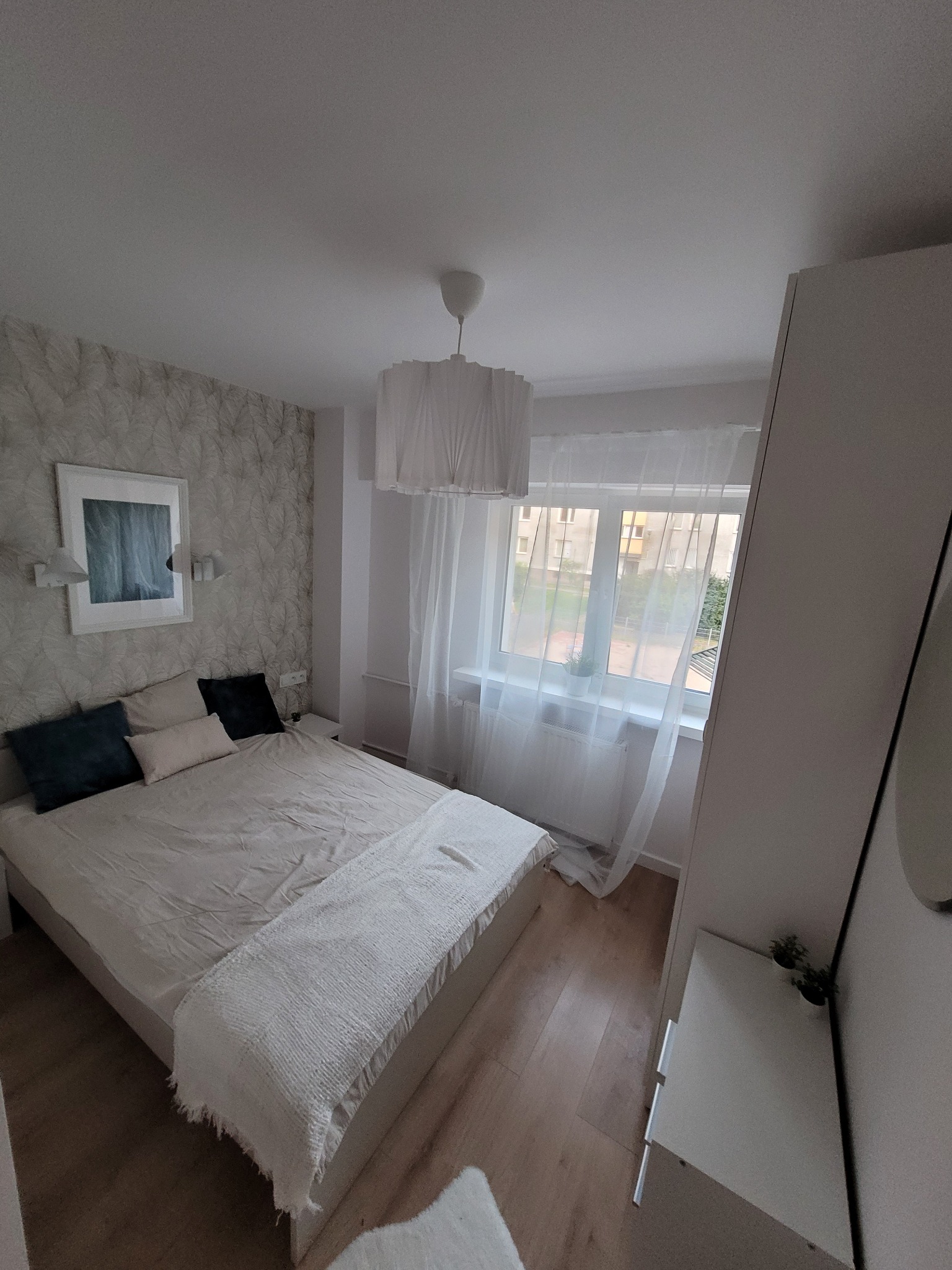 Jasna sypialnia z łóżkiem, wzorzystą tapetą i białymi zasłonami w oknie, widok na budynki z zewnątrz.