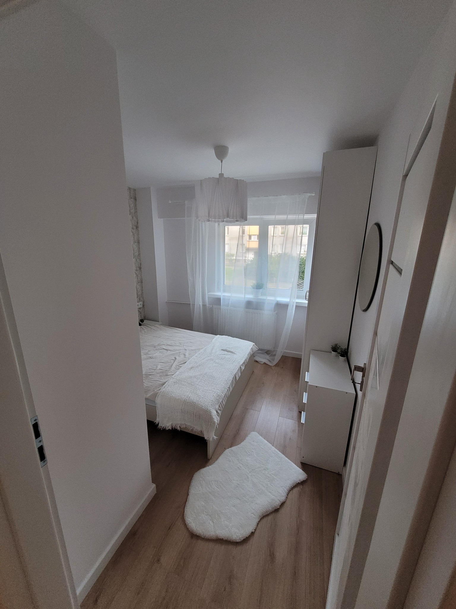 Jasna sypialnia z drewnianą podłogą, białym łóżkiem z narzutą, puszystym dywanikiem i szafką nocną pod oknem z białymi zasłonami.