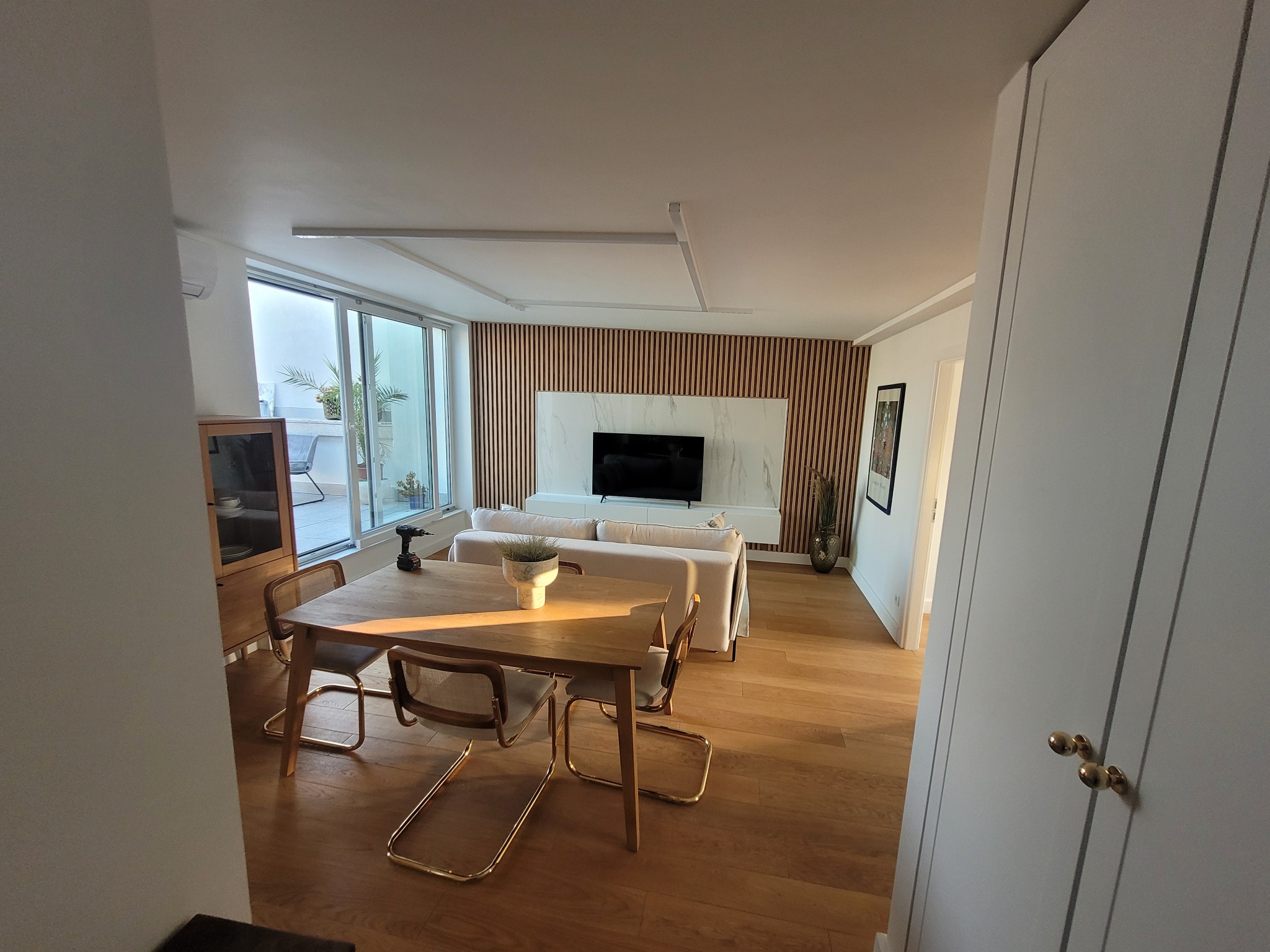 Jasne, przestronne wnętrze mieszkania z drewnianą podłogą i panelami na ścianie za telewizorem. Na pierwszym planie stół z czterema krzesłami, na którym leży wiertarka akumulatorowa.