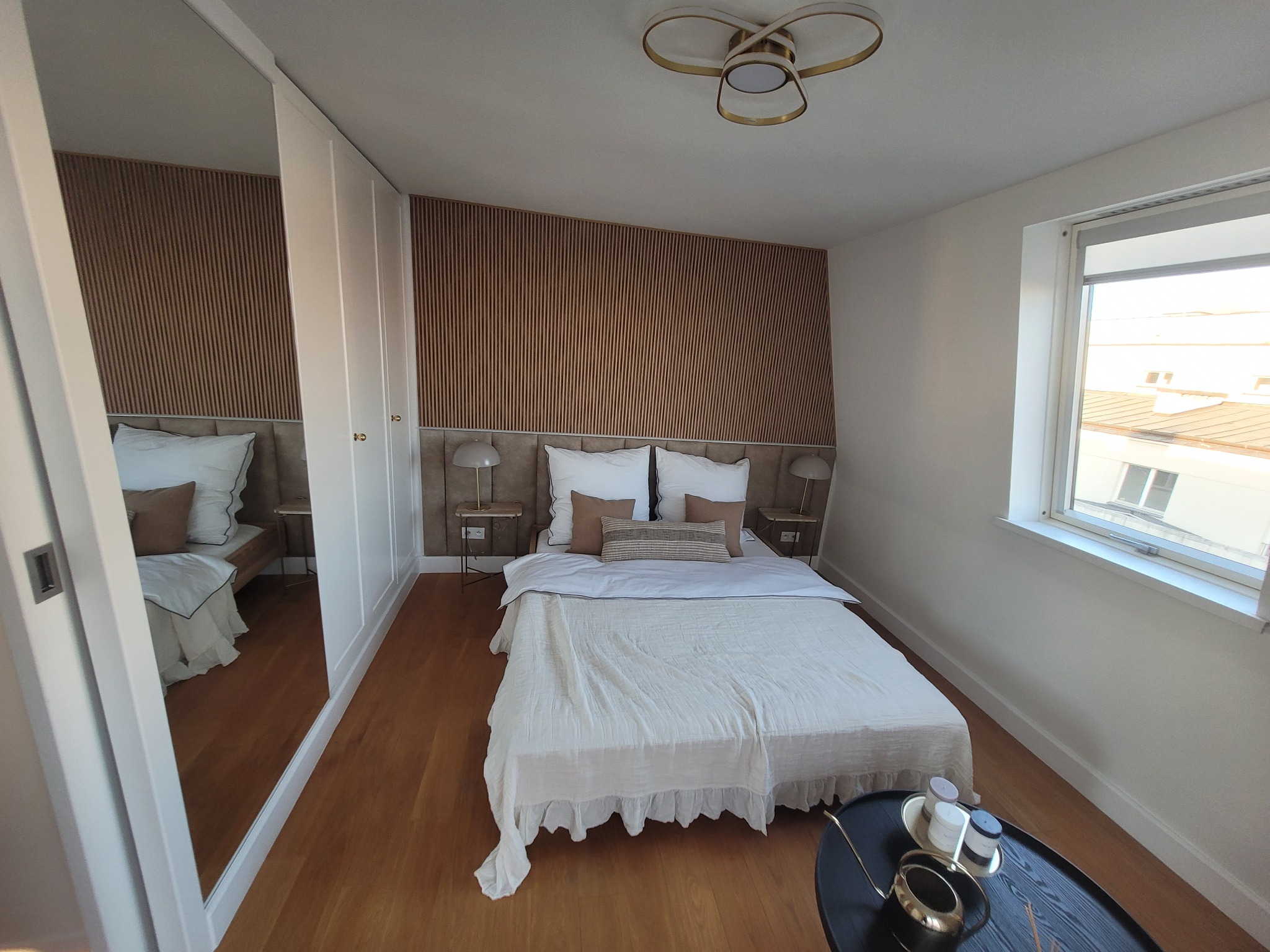 Jasna sypialnia z drewnianą boazerią na ścianie za łóżkiem, parkietem na podłodze, białymi szafami wnękowymi z lustrem i stolikiem kawowym z dekoracjami obok okna.