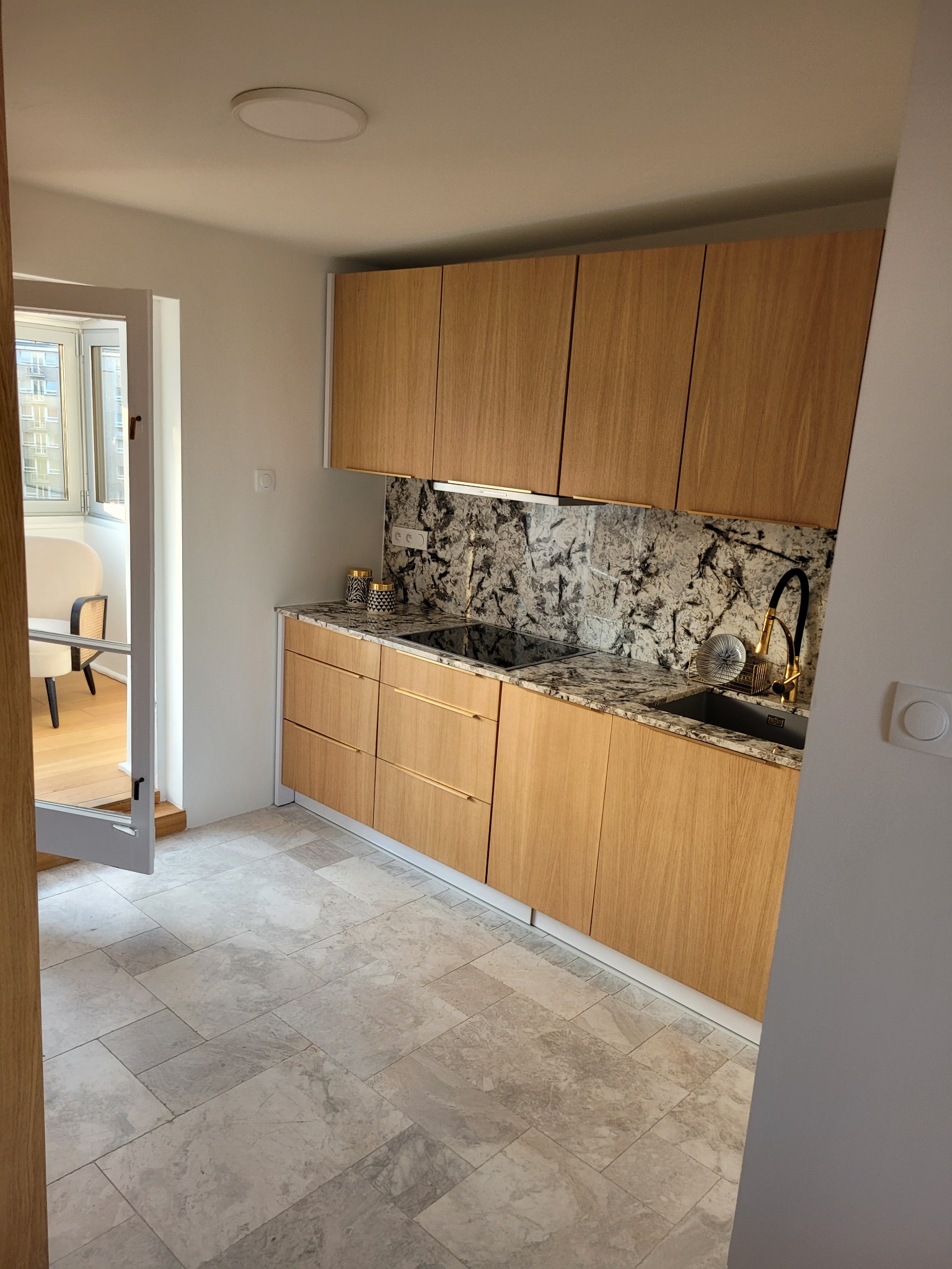 Jasna kuchnia z drewnianymi szafkami, marmurowym blatem i złotą armaturą, widok na balkon z krzesłem przez otwarte drzwi balkonowe.