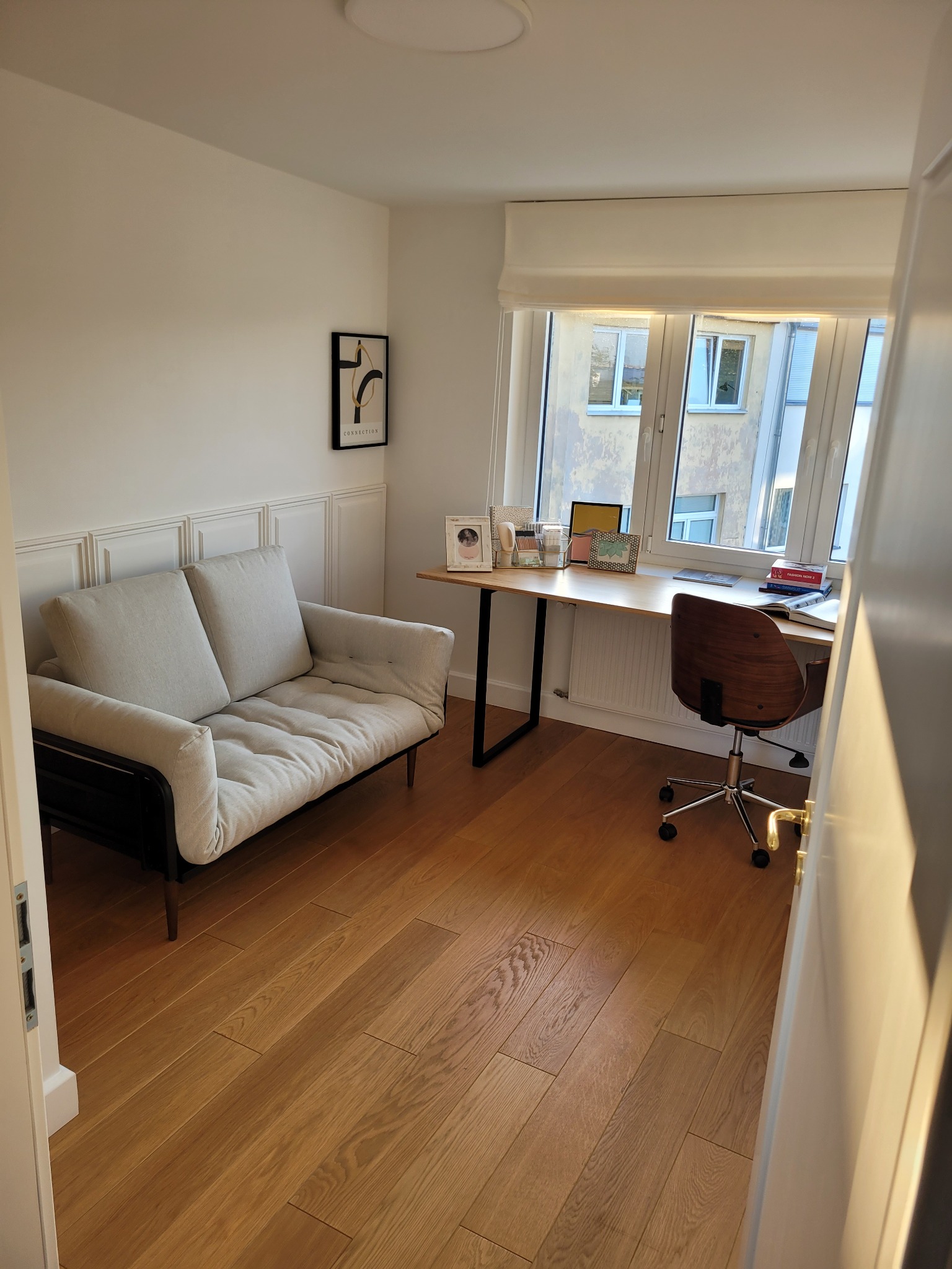 Jasne panele podłogowe w pokoju z sofą, biurkiem przy oknie i dekoracyjną boazerią ścienną.