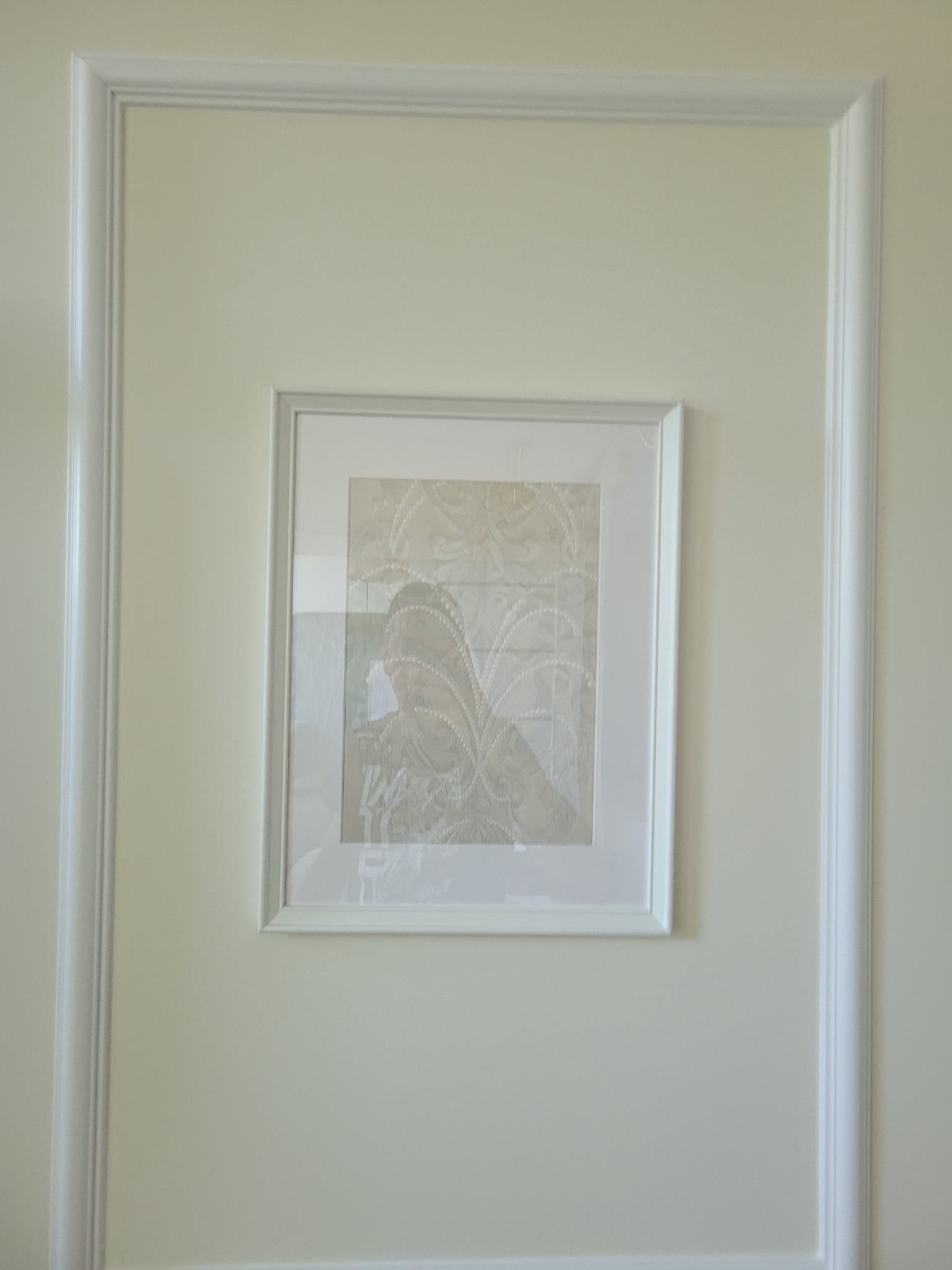 Ściana w kolorze złamanej bieli z dekoracyjną ramą i zawieszonym obrazem w białej ramce, przedstawiającym abstrakcyjny wzór w odcieniach beżu i szarości.