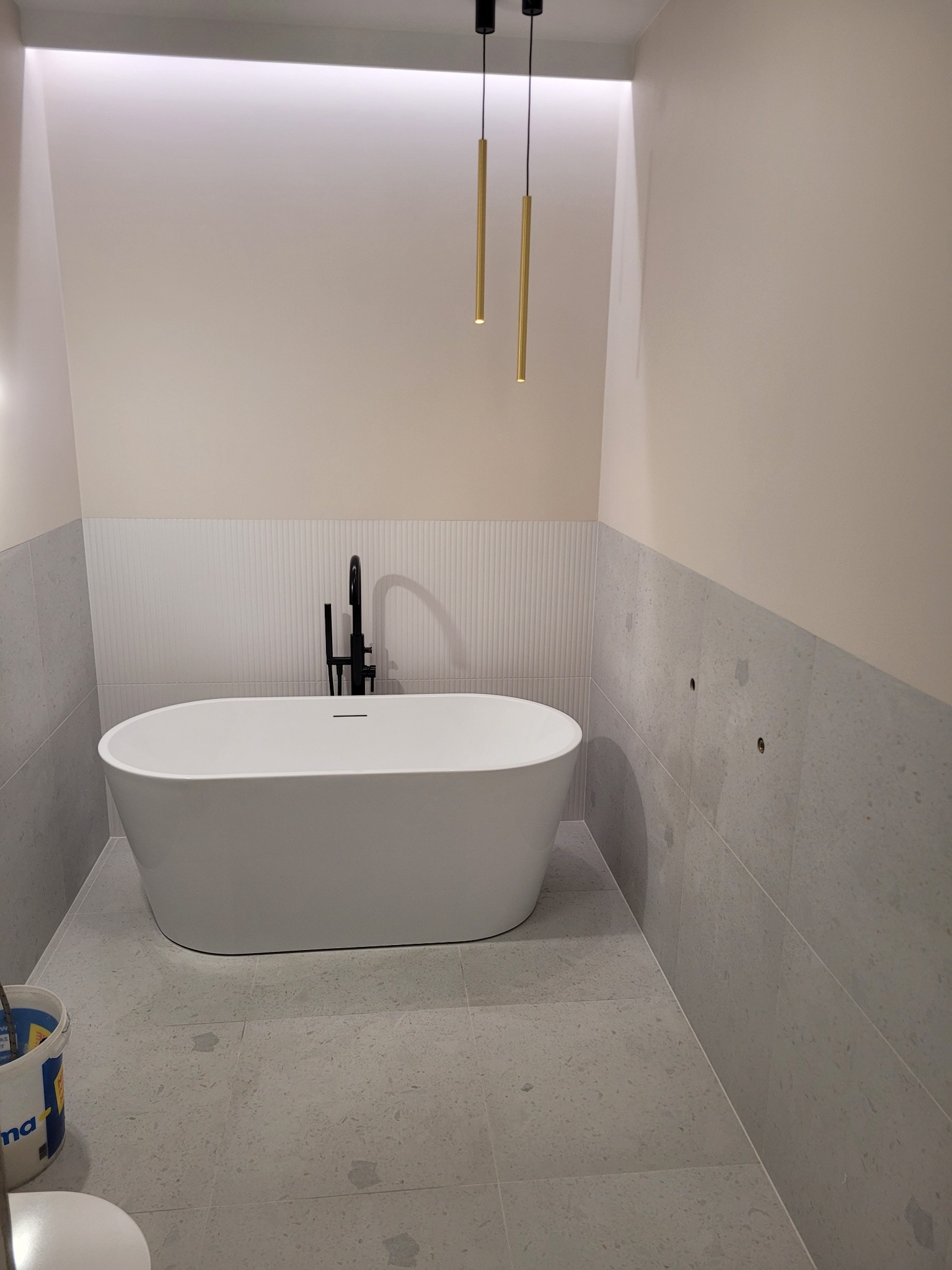 Nowoczesna łazienka z wolnostojącą białą wanną, czarną armaturą i szarymi płytkami imitującymi lastryko na podłodze i ścianach do połowy wysokości, oświetlona wiszącymi złotymi lampami.