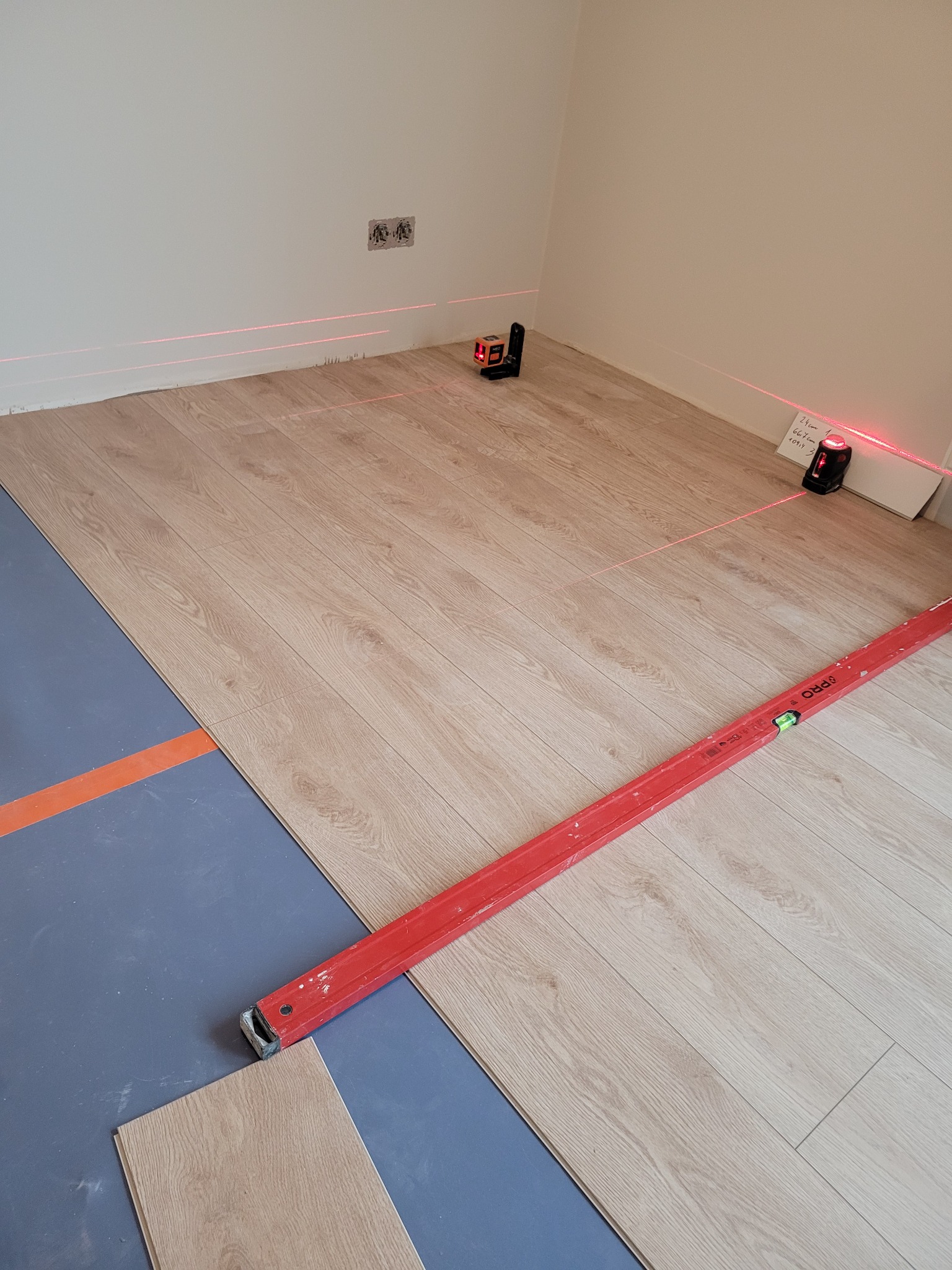Fragment pokoju w trakcie montażu podłogi z paneli; widoczne panele ułożone na podkładzie, poziomica, laser krzyżowy wyznaczający linie oraz niezamontowane panele.