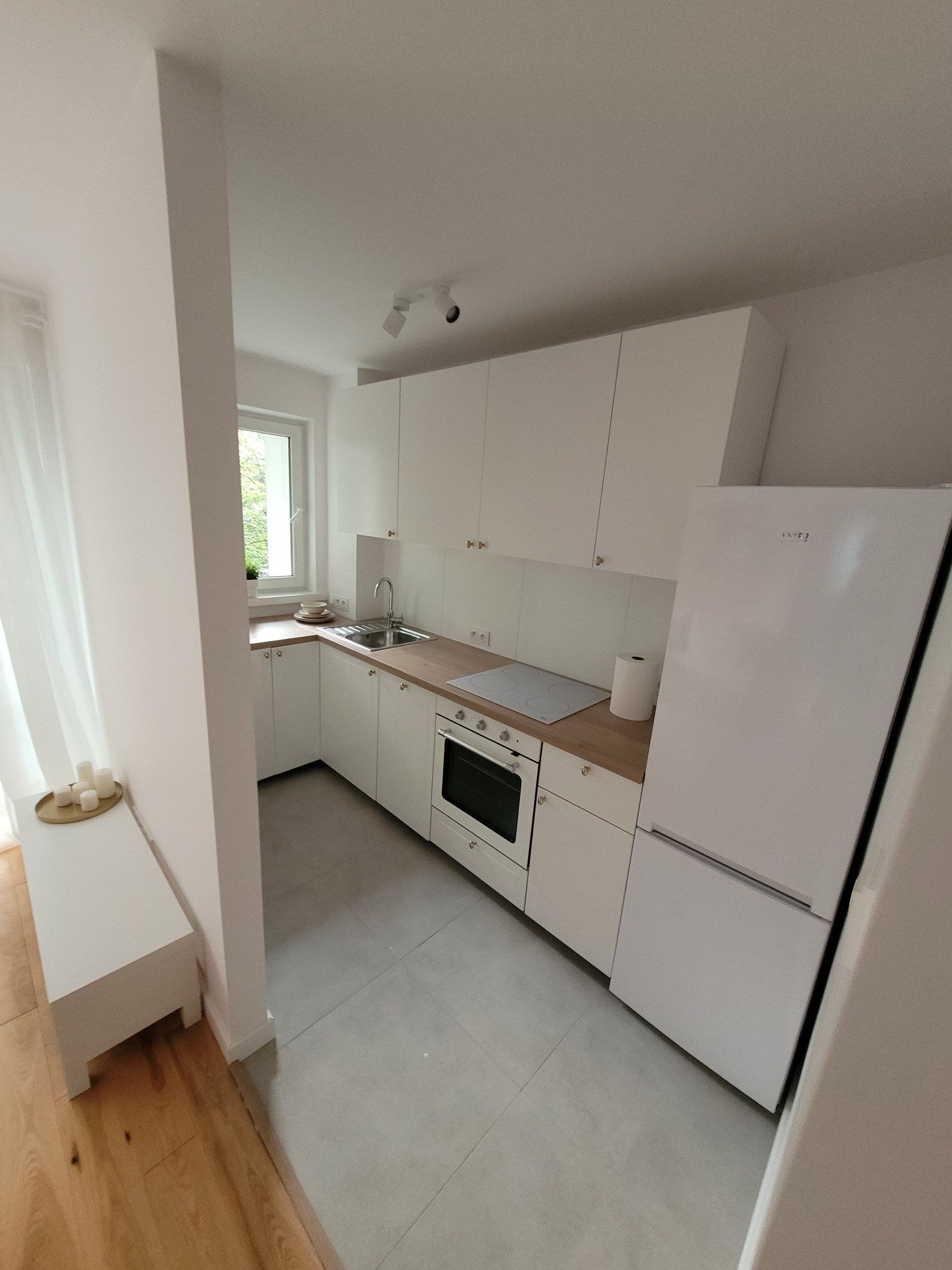 Jasna kuchnia z białymi szafkami, drewnianym blatem i szarą podłogą, widok z perspektywy wejścia, z dekoracyjnymi świeczkami na parapecie.