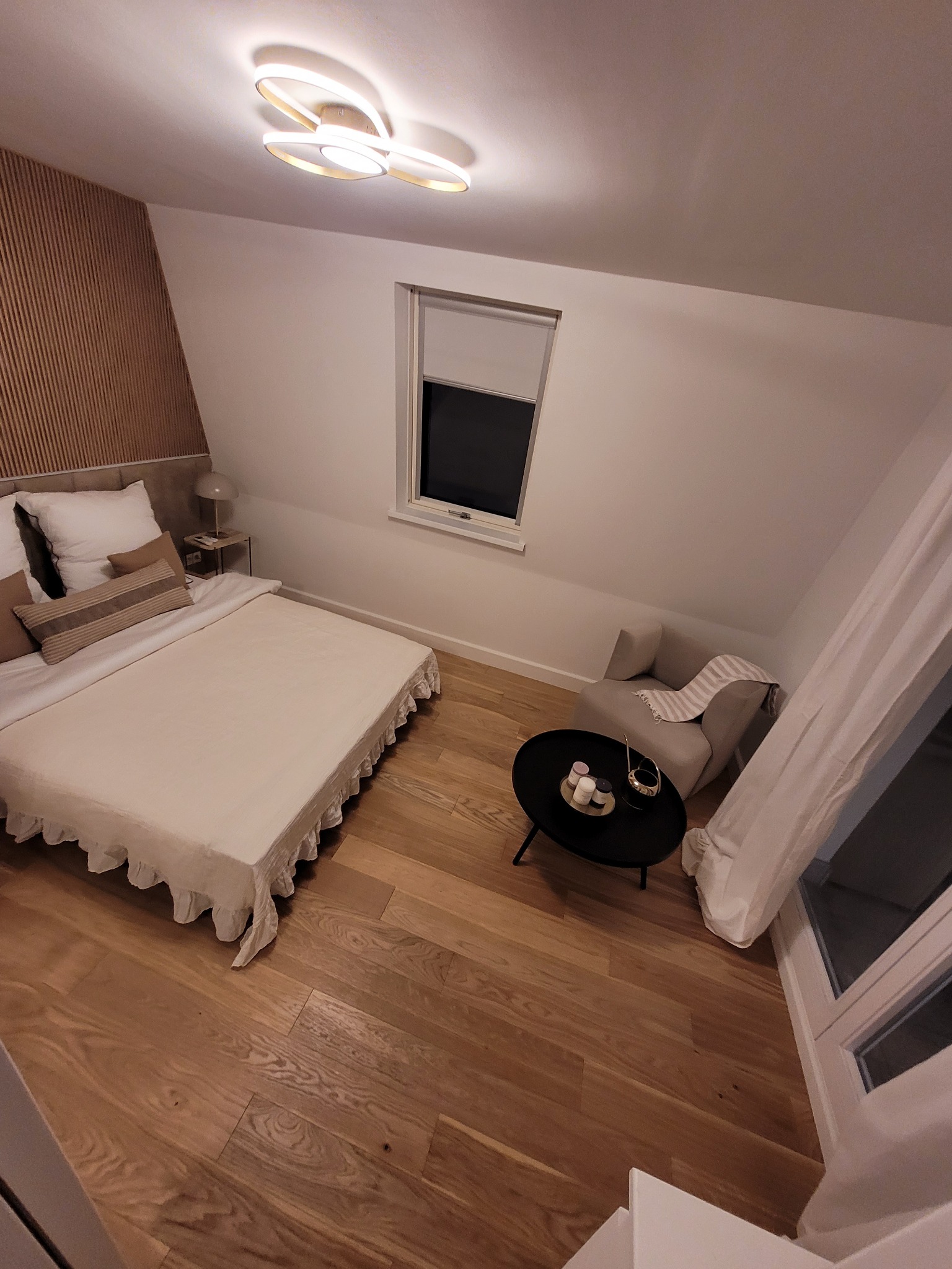 Jasna sypialnia na poddaszu z drewnianą podłogą, łóżkiem z beżową narzutą, stolikiem kawowym i krzesłem, oświetlona nowoczesną lampą sufitową.