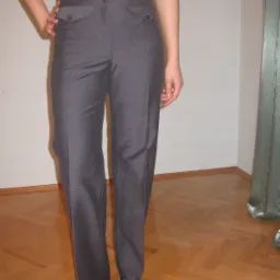 Prezentacja szarych, damskich spodni wizytowych z wysokim stanem i kieszeniami, w zestawieniu z białą koszulą i czarnymi butami na obcasie z ozdobnymi kokardkami, na tle parkietu i białej ściany.