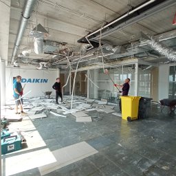 RB-Roman Bratasiuk - Wnętrze biura w trakcie demontażu, widoczni trzej pracownicy w trakcie pracy, gruz i śmieci na podłodze, żółty kontener na odpady, logo firmy Daikin na ścianie w tle.