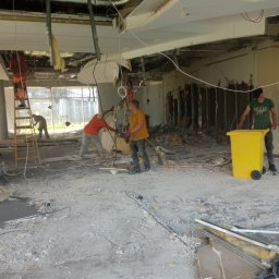 RB-Roman Bratasiuk - Wnętrze w trakcie rozbiórki: pracownicy usuwają elementy ścian i sufitu, widoczny gruz i żółty kontener na odpady.