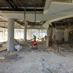 RB-Roman Bratasiuk - Wnętrze budynku w trakcie rozbiórki, widoczne zniszczone elementy sufitu, ścian i podłogi, oraz pracownicy w odblaskowych ubraniach usuwający gruz, z widocznymi instalacjami i rurami pod sufitem.