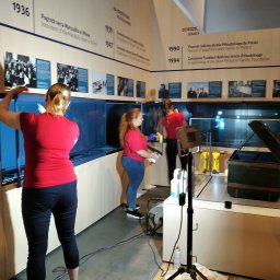 RB-Roman Bratasiuk - Pracownice w czerwonych koszulkach czyszczą ekspozycję w Muzeum Józefa Piłsudskiego w Sulejówku, widoczne gabloty z eksponatami i tablice informacyjne z datami i opisami historycznymi.