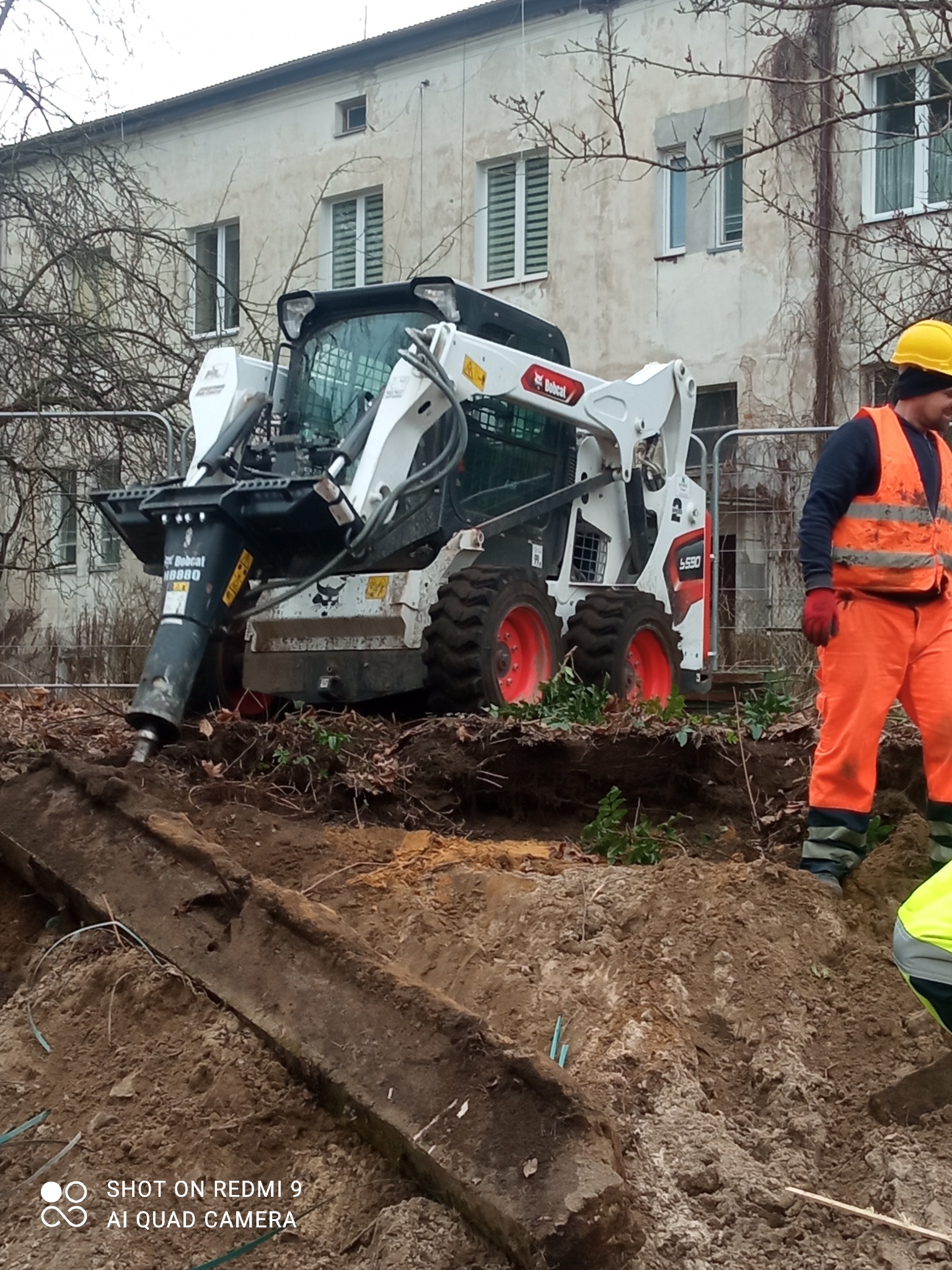 Biała miniładowarka Bobcat S530 z młotem hydraulicznym HB880 rozbija betonowy element w ziemi, obok pracownik w pomarańczowym kombinezonie i żółtym kasku, w tle budynek z szarą elewacją.