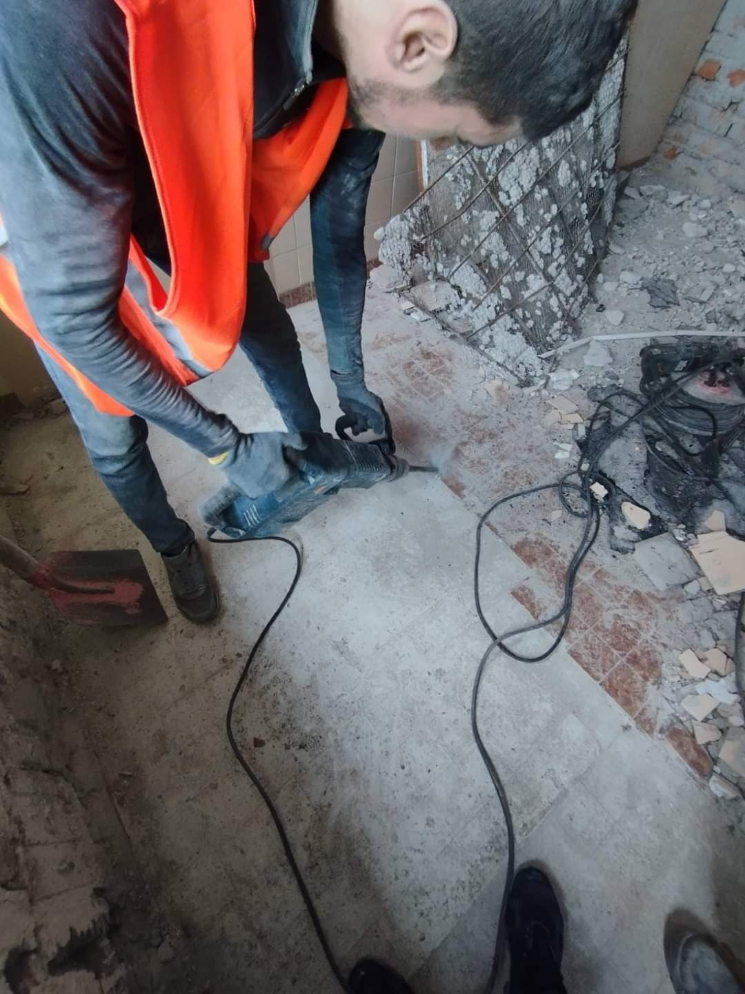Pracownik w pomarańczowej kamizelce używa młota udarowego do skuwania płytek podłogowych w trakcie remontu łazienki, widoczne gruz i kabel zasilający.