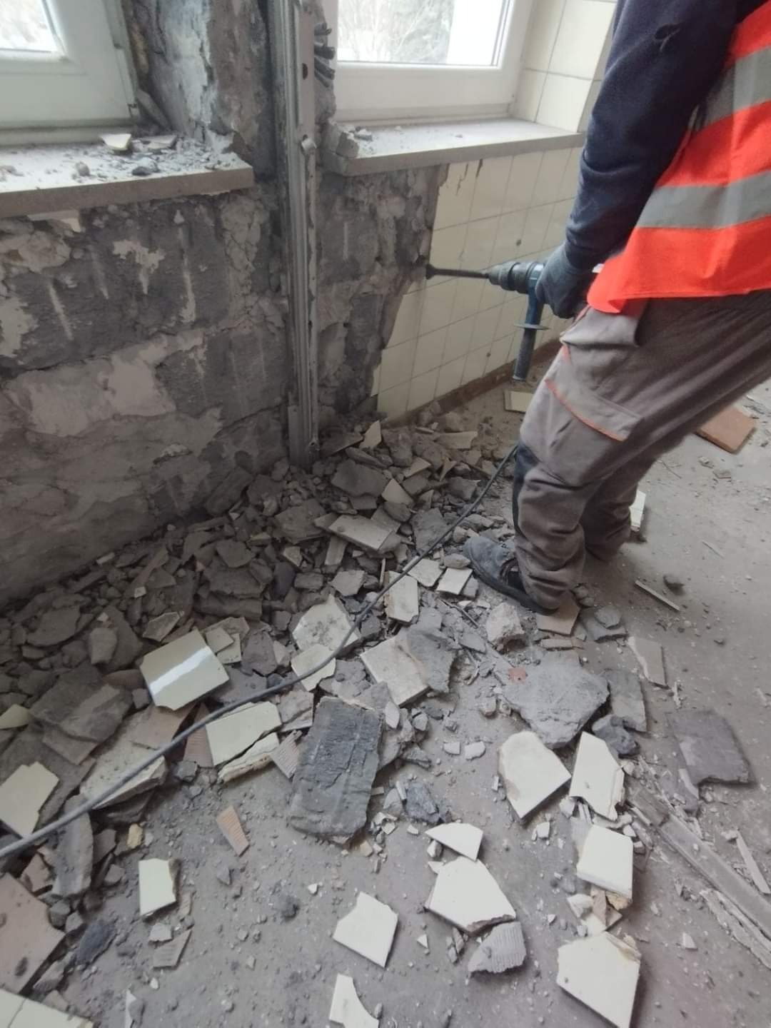 Pracownik w kamizelce odblaskowej używa wiertarki udarowej do usuwania płytek ściennych w pobliżu okna, z gruzem na podłodze.