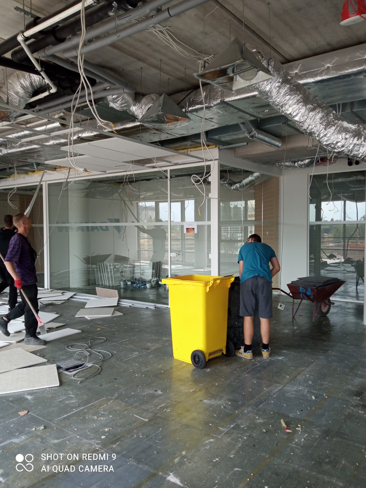 Wnętrze biura w trakcie demontażu: widoczne usunięte płyty sufitowe, resztki instalacji, pracownicy z narzędziami i żółty kontener na odpady.