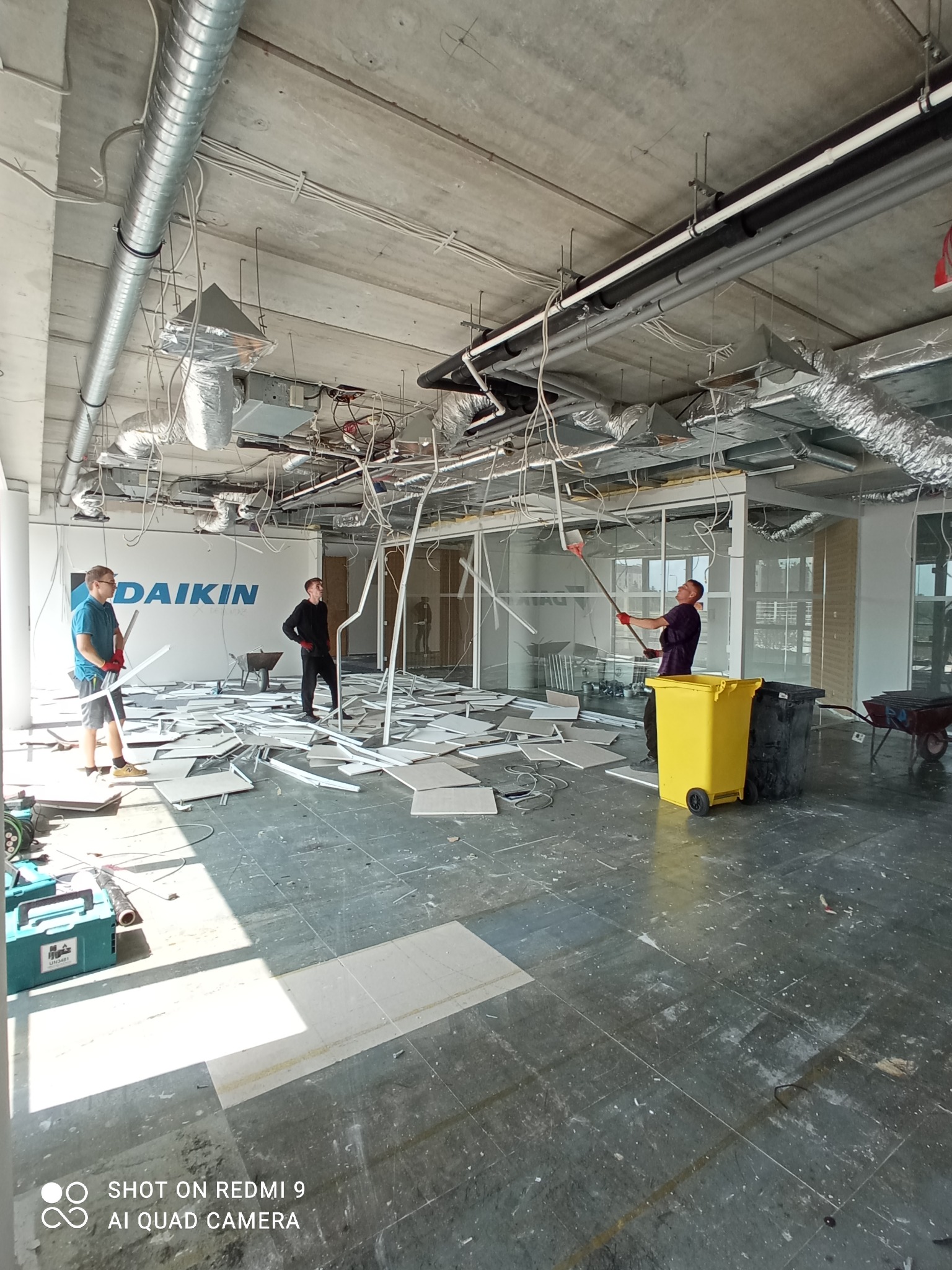 Wnętrze biura w trakcie demontażu, widoczni trzej pracownicy w trakcie pracy, gruz i śmieci na podłodze, żółty kontener na odpady, logo firmy Daikin na ścianie w tle.