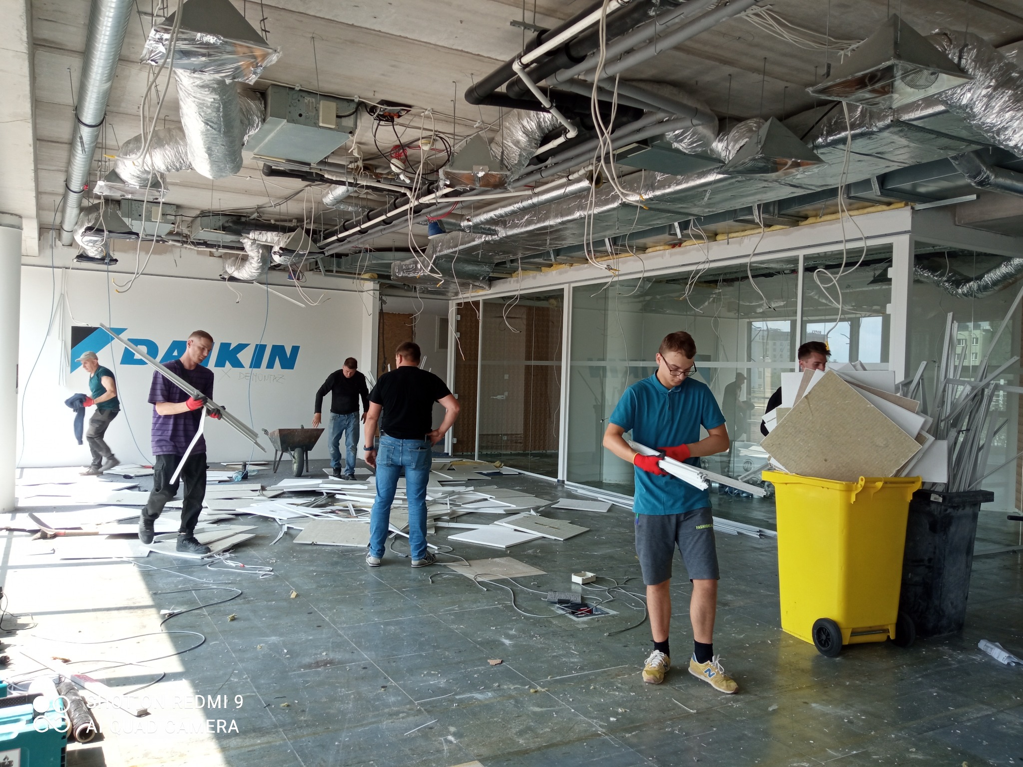 Demontaż instalacji w biurze: pracownicy w trakcie usuwania elementów, widoczne przewody, fragmenty sufitu, żółty kontener na odpady, logo Daikin na ścianie w tle.