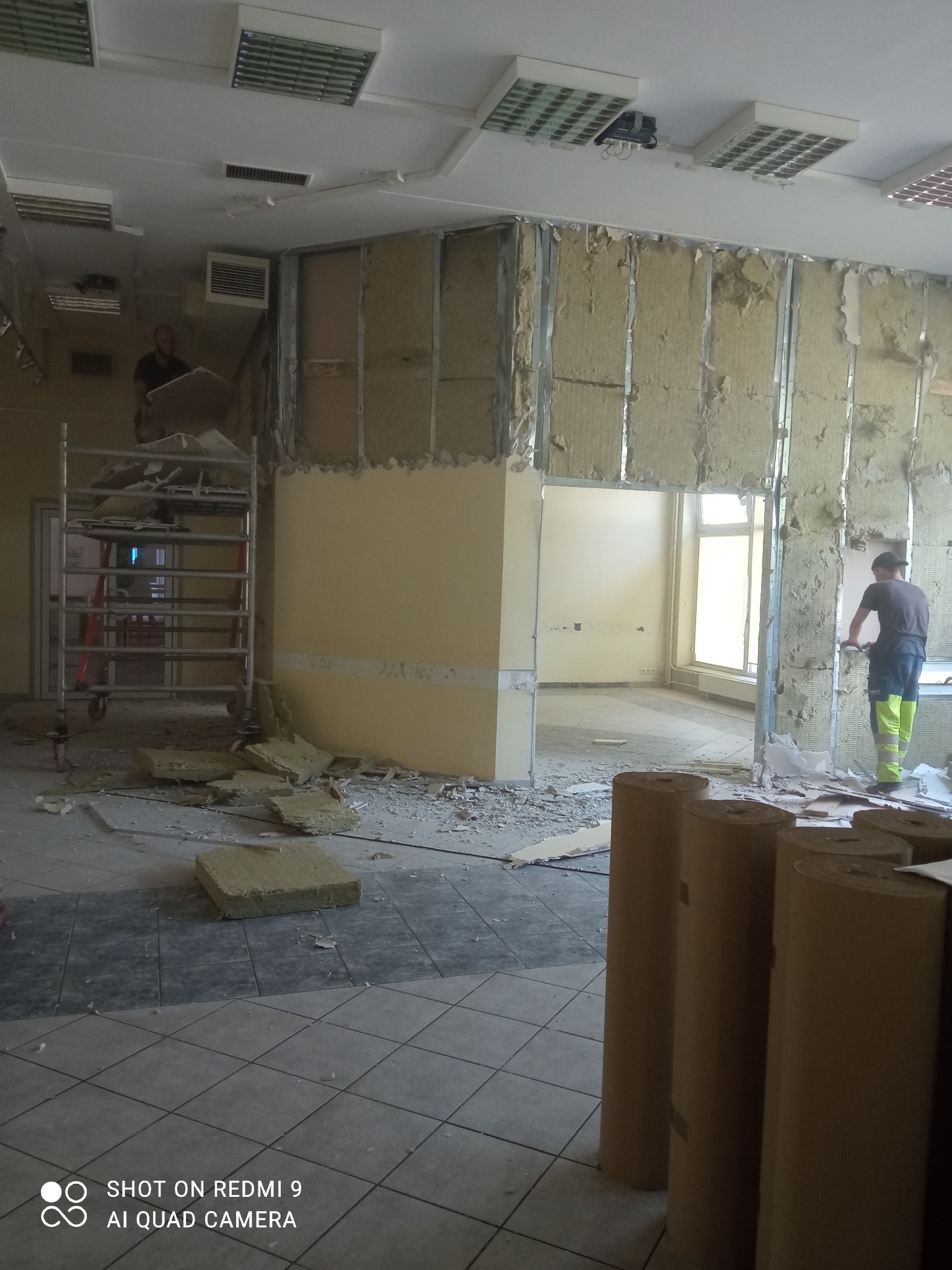 Demontaż ścian działowych w biurze: pracownik na rusztowaniu przenosi płyty, drugi przy ścianie z izolacją z wełny mineralnej, widoczne rolki materiału izolacyjnego na podłodze z płytek.