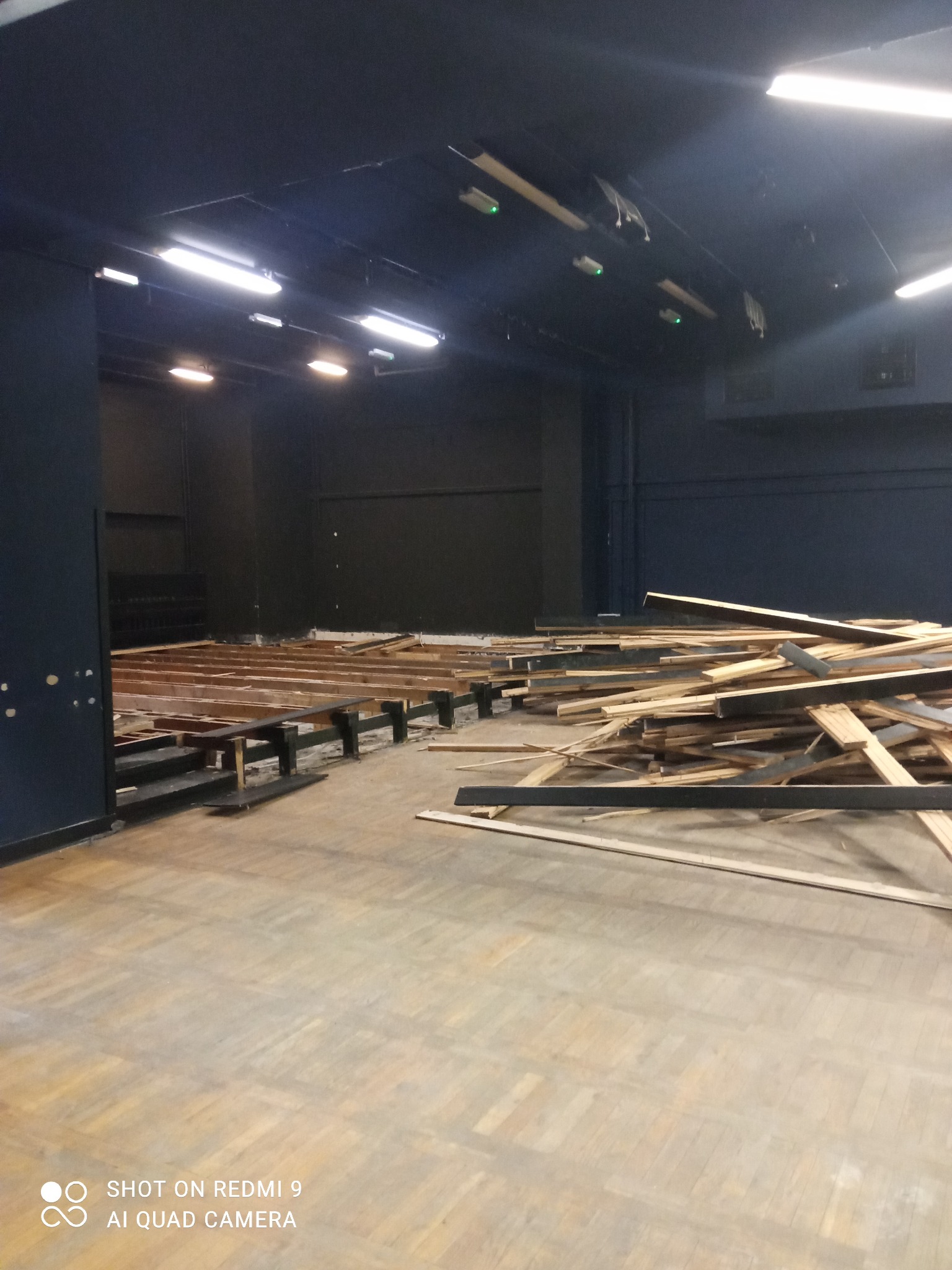 Wnętrze w trakcie demontażu drewnianej konstrukcji, widoczne rozłożone elementy i sterta desek na parkiecie, czarne ściany i sufit z oświetleniem.