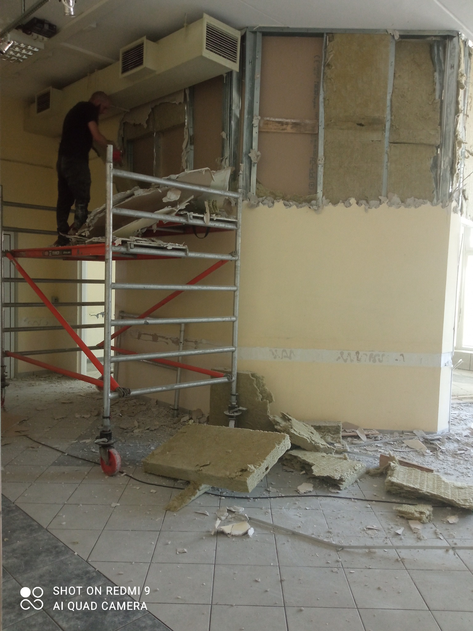 Pracownik na rusztowaniu demontuje ścianę z widoczną izolacją z wełny mineralnej, gruz na podłodze, widoczne instalacje wentylacyjne pod sufitem.