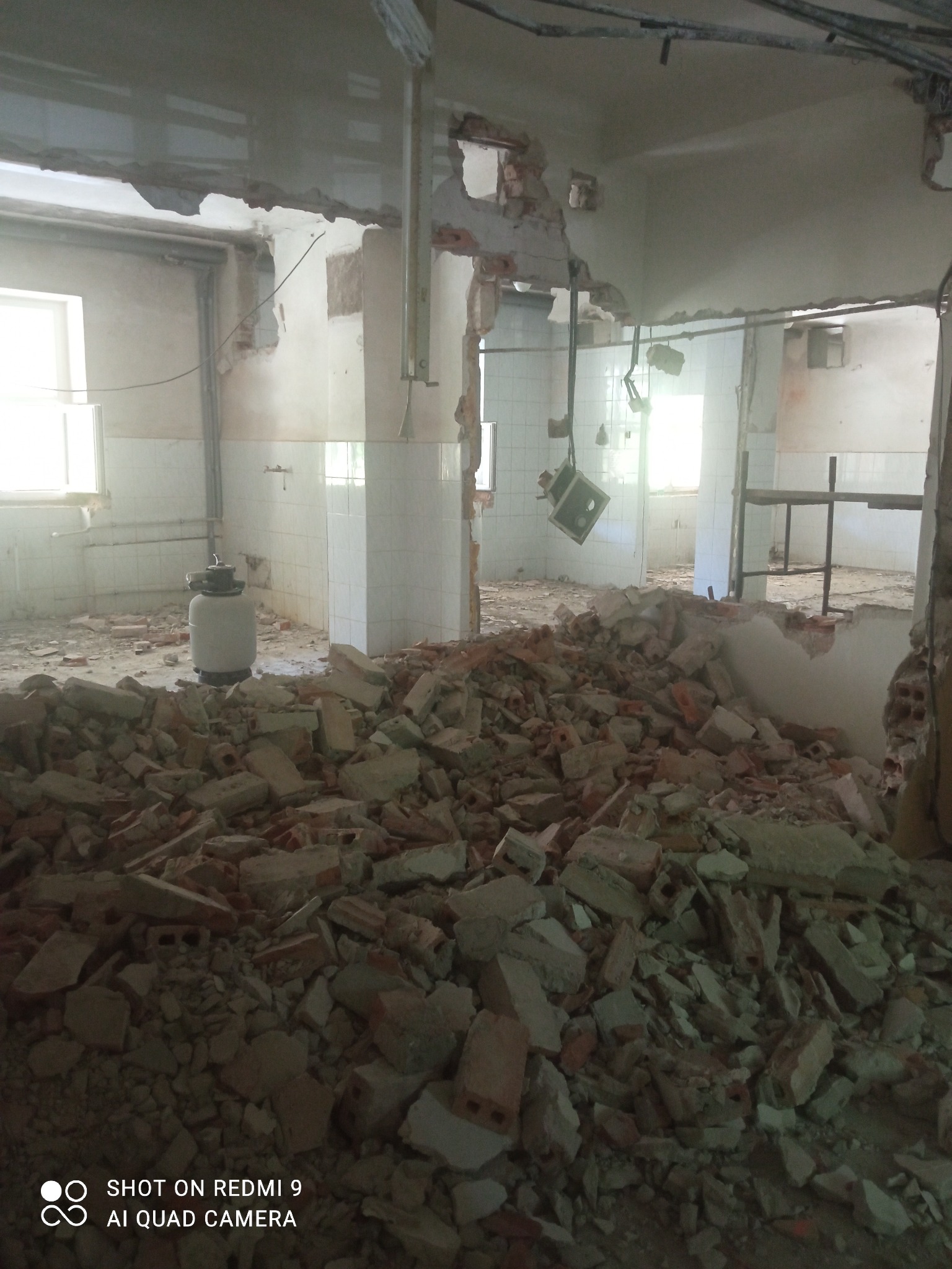 Wnętrze po wyburzeniu ściany działowej, widoczny gruz ceglany i fragmenty instalacji, białe kafelki na ścianach w tle, światło dzienne wpadające przez okna.