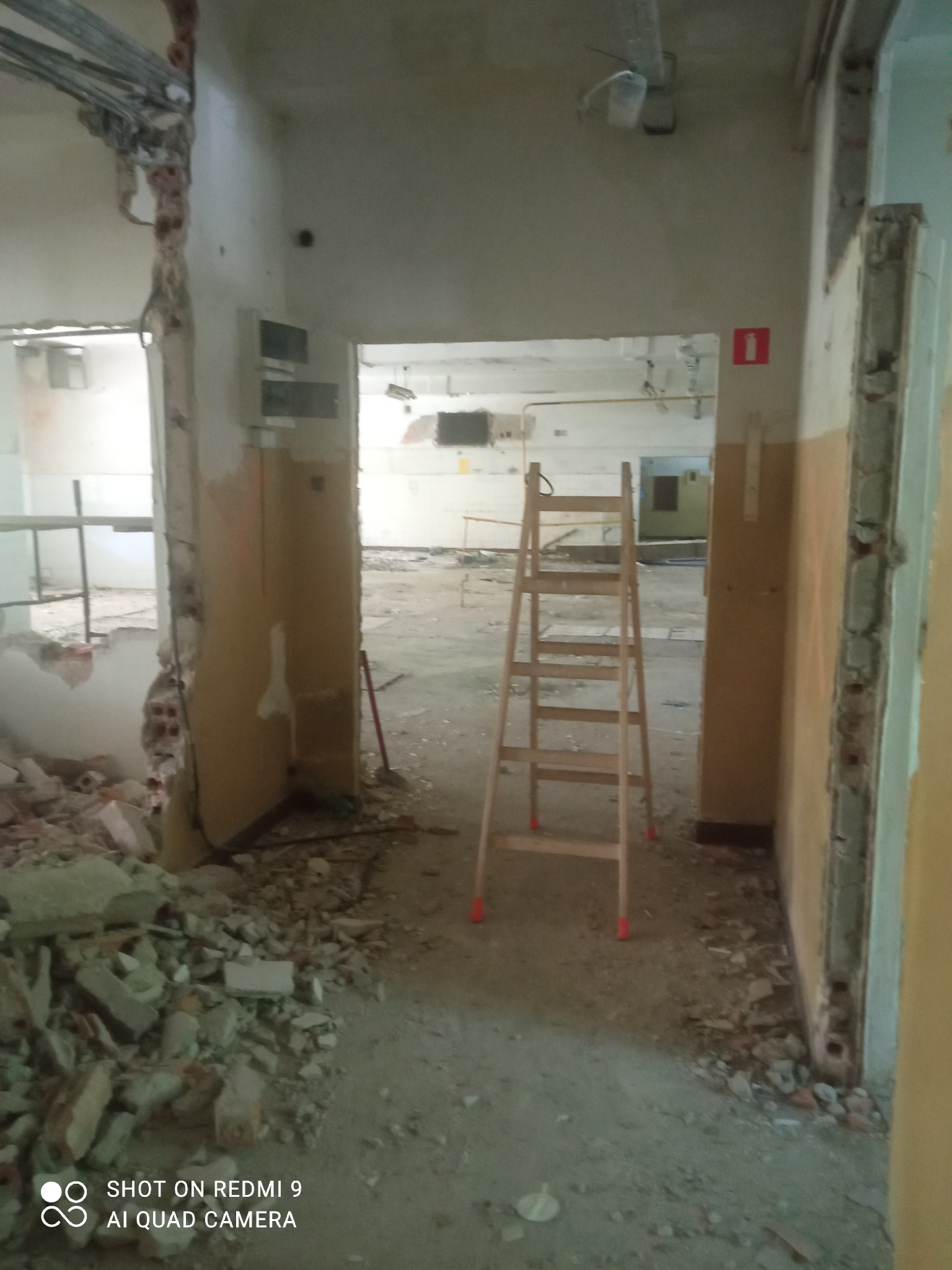 Wnętrze budynku w trakcie rozbiórki, widoczny gruz, uszkodzone ściany i drabina w przejściu.